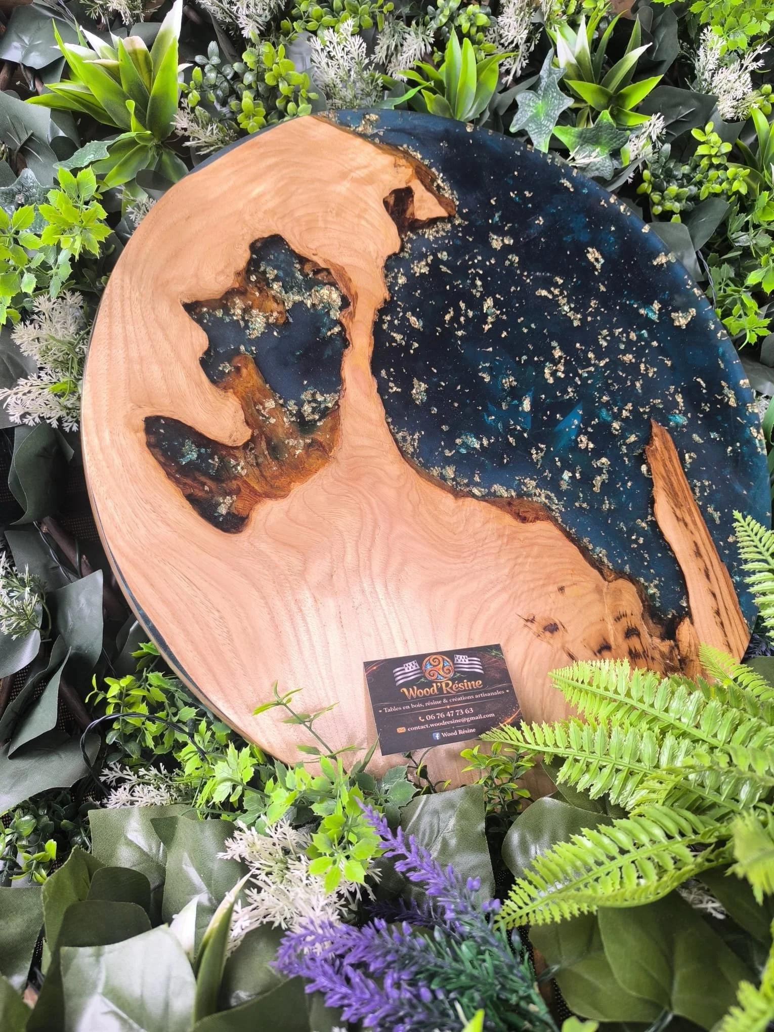 Table en bois avec des incrustations de résine bleue et dorée, entourée de plantes vertes et de fleurs.