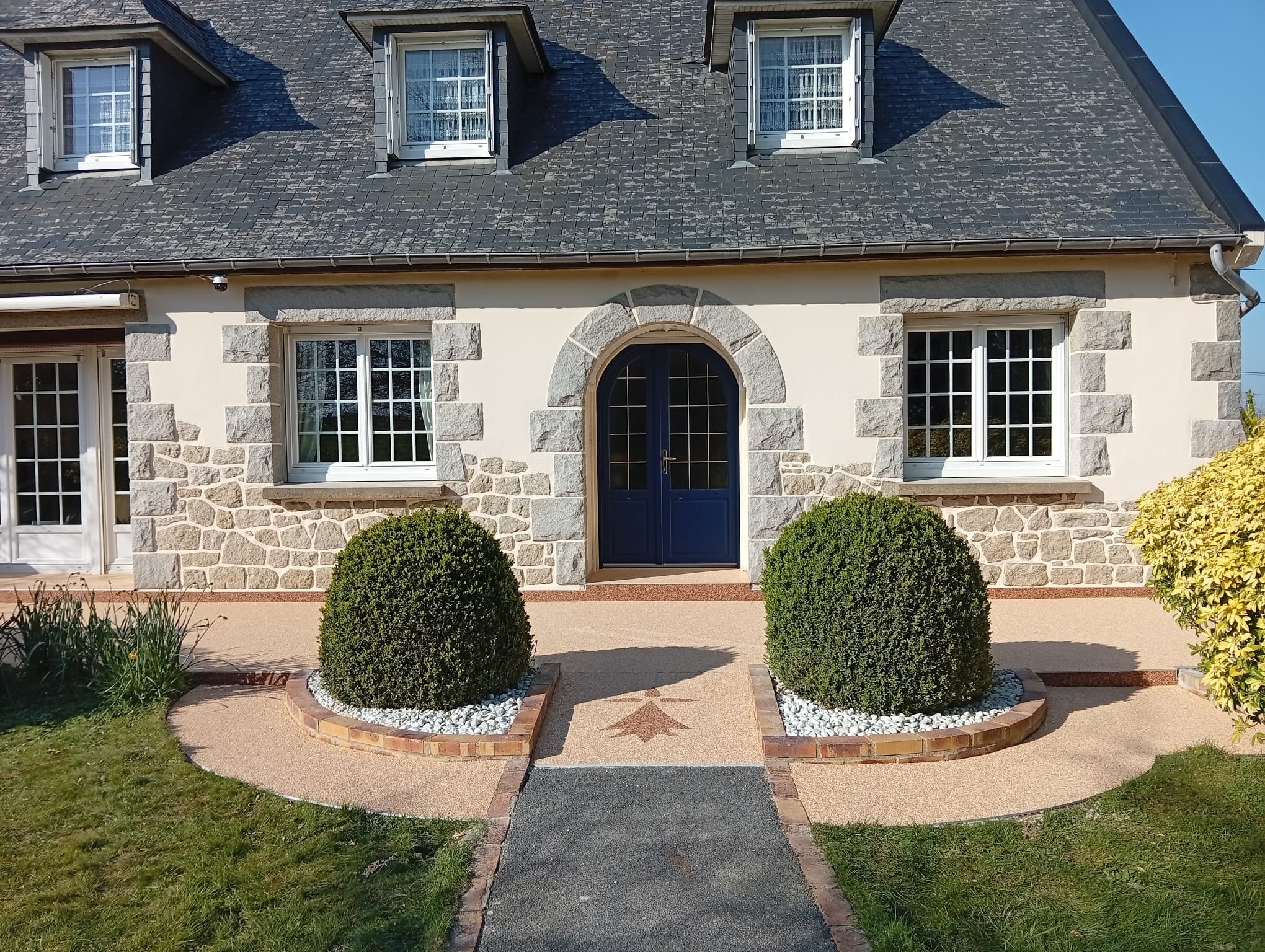 Maison avec façade en pierre, fenêtres à volets gris, porte bleue dans une arche, jardinières avec buissons arrondis, allée en béton et pavés, ciel clair.