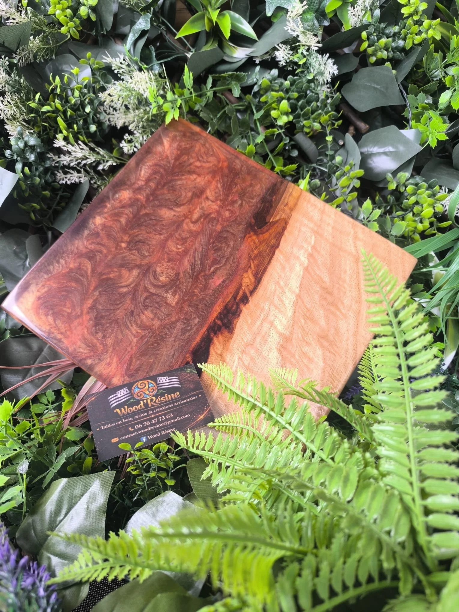 Une planche en bois résine avec deux styles différents, entourée de diverses plantes vertes et un badge de contact pour Wood Résine.