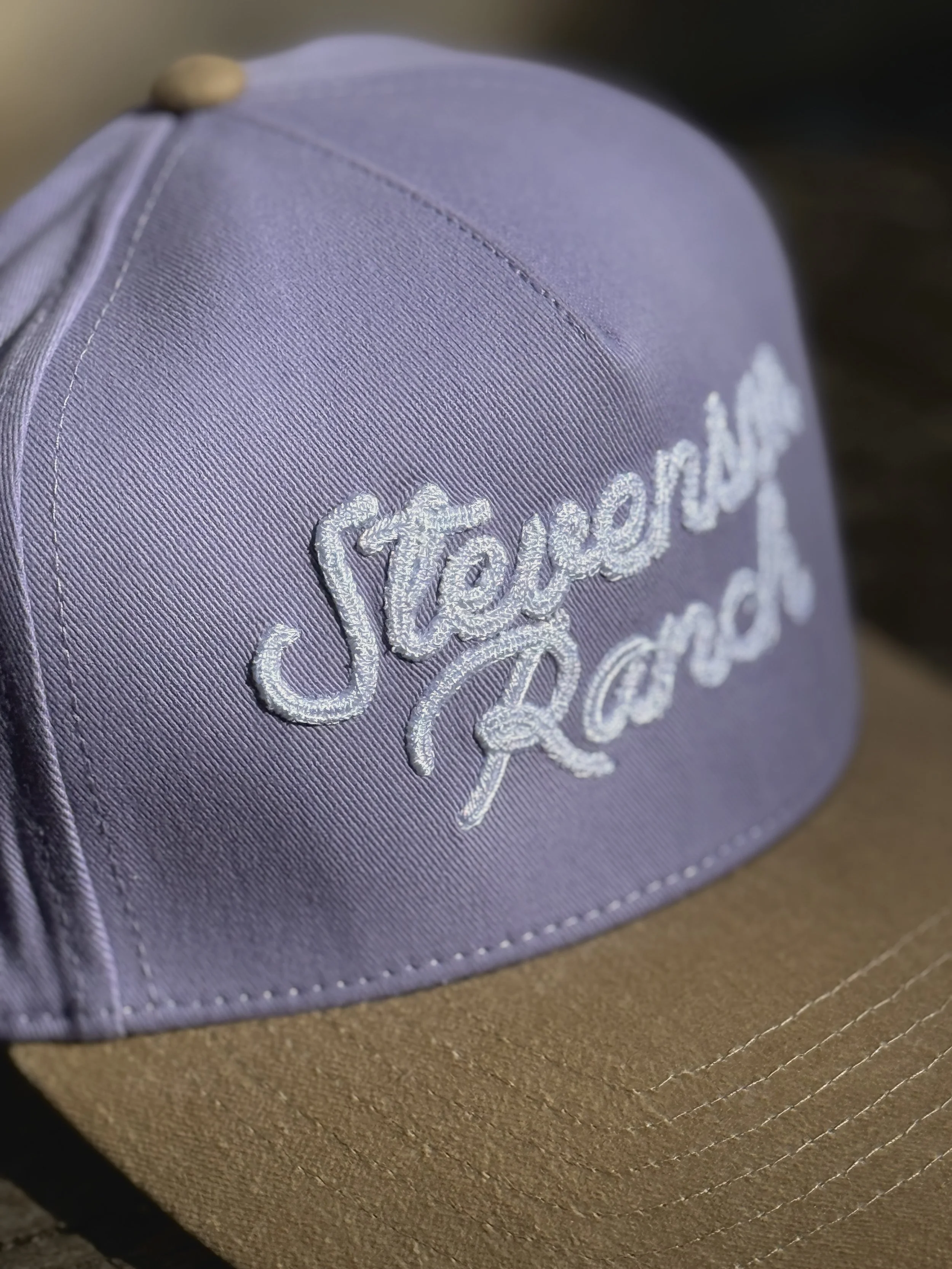 Close-up of a lavender baseball cap with the white embroidered logo "Vander" on the front, resting on a brown surface.
