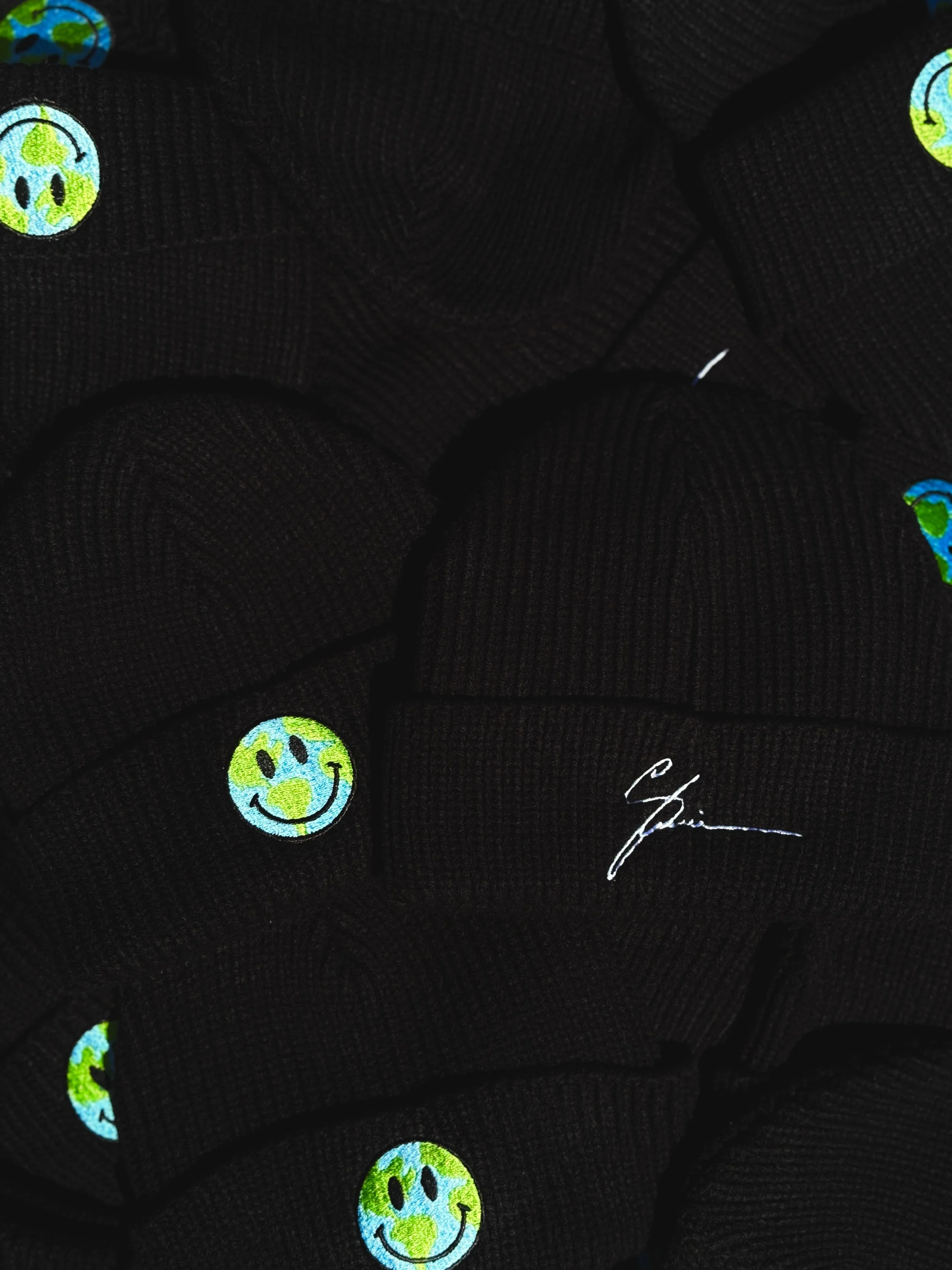 Close-up of black fabric with embroidered smiley face icons with Earth's continents as the face, and a white signature or name embroidered near the bottom right.