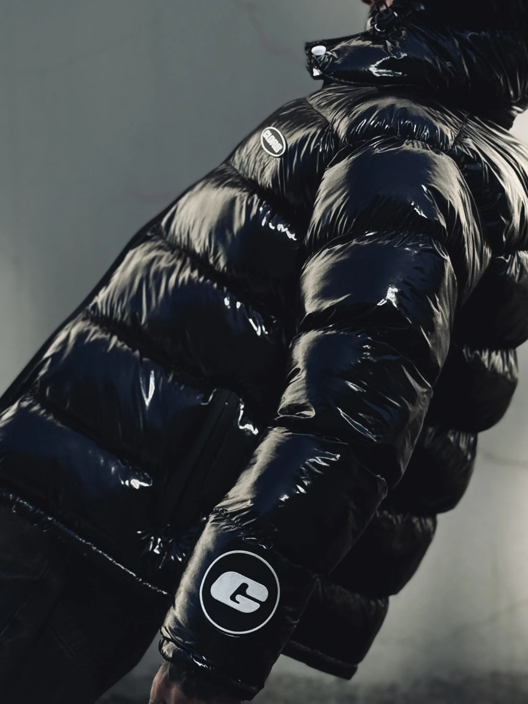 Close-up of a shiny black puffer jacket with logos, worn by a person standing against a blurred gray background.