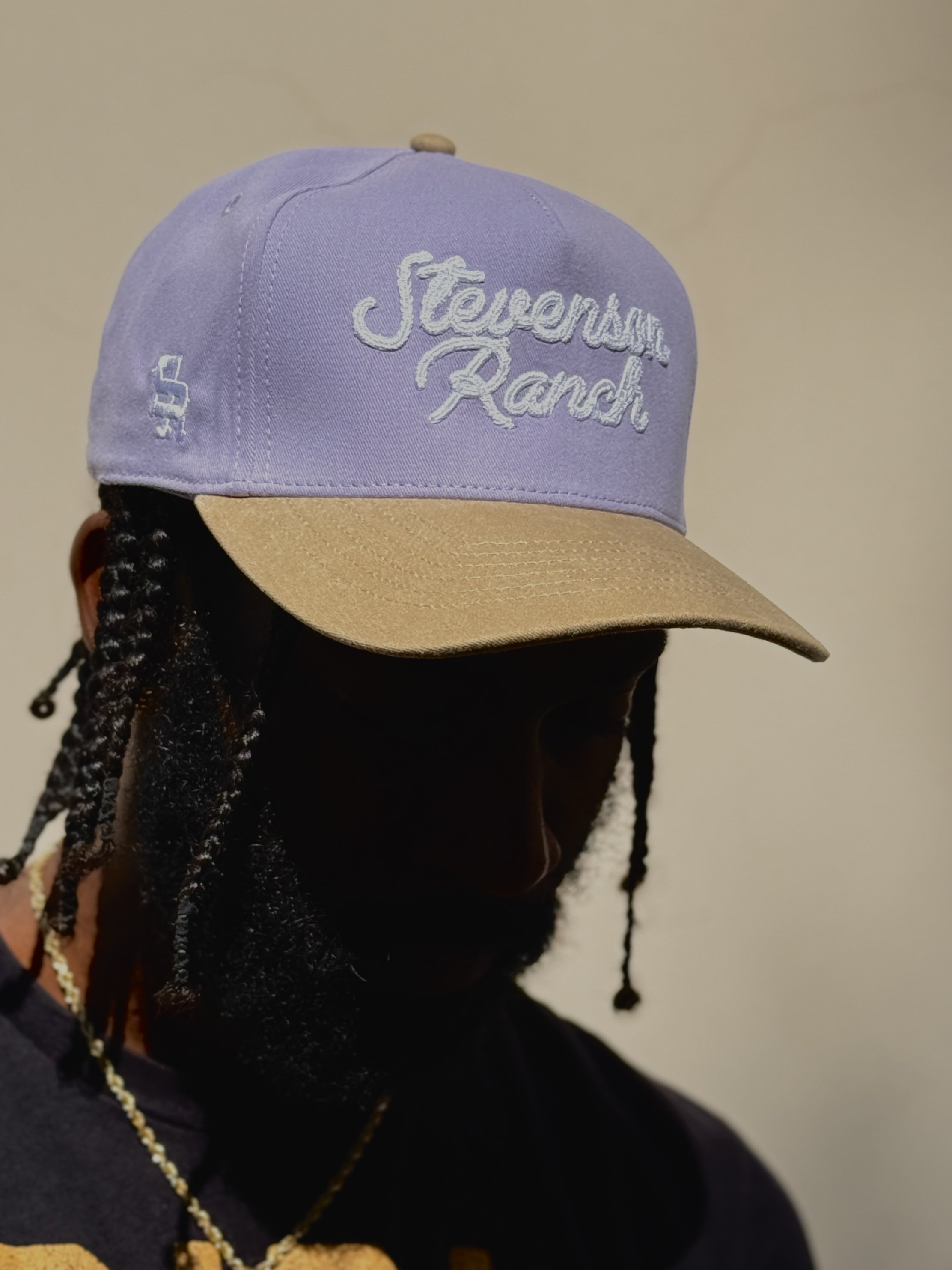 Close-up of a person's head wearing a lavender baseball cap with 'Steve'em Ranch' embroidered in white on the front, and a tan brim. The person's face is not visible, and they have braids. The background is plain and neutral.