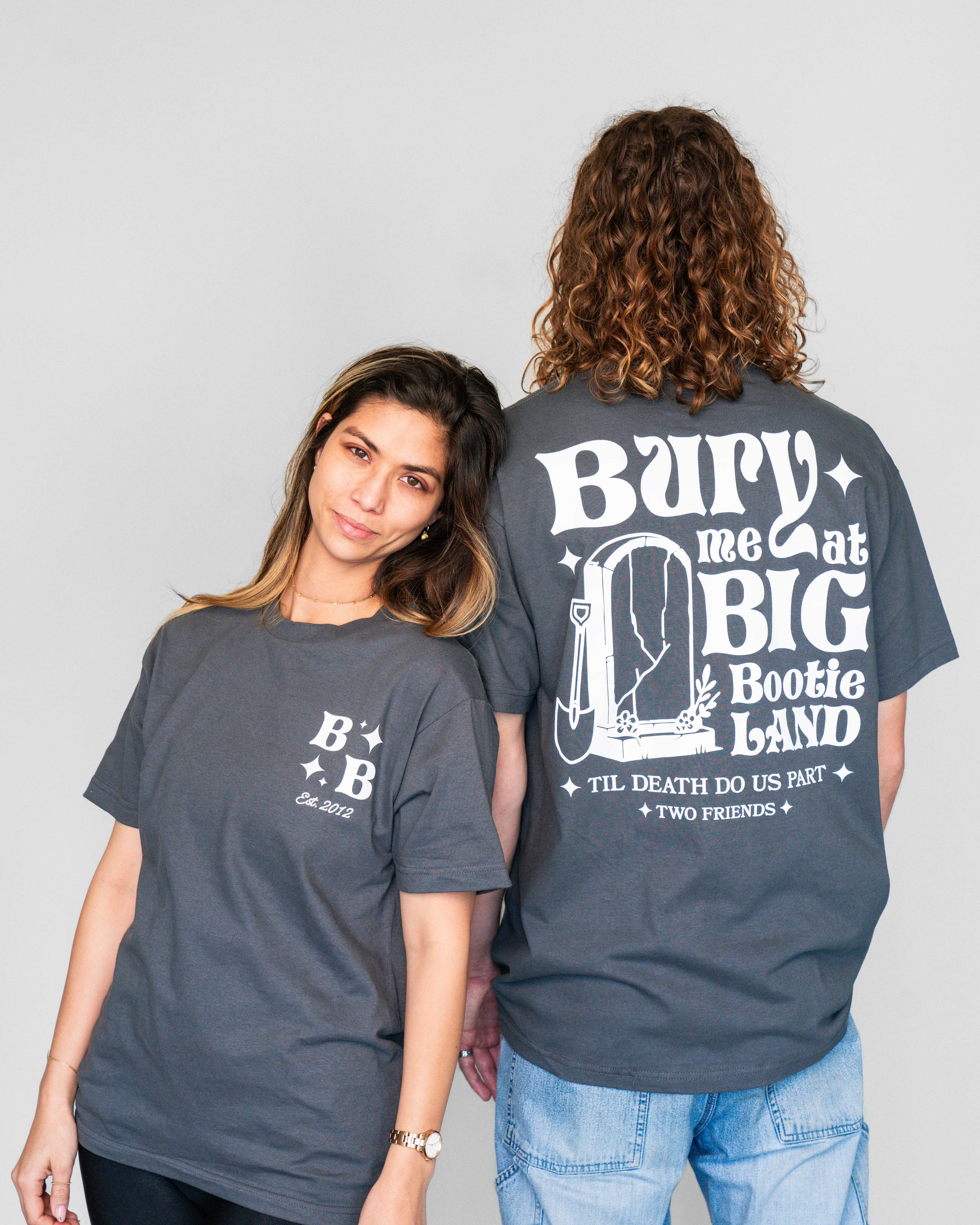 Two women wearing matching dark gray t-shirts standing side by side against a plain gray background. The woman on the left has long brown hair, is smiling subtly, and is wearing a gold watch and jewelry. The woman on the right has curly brown hair, b