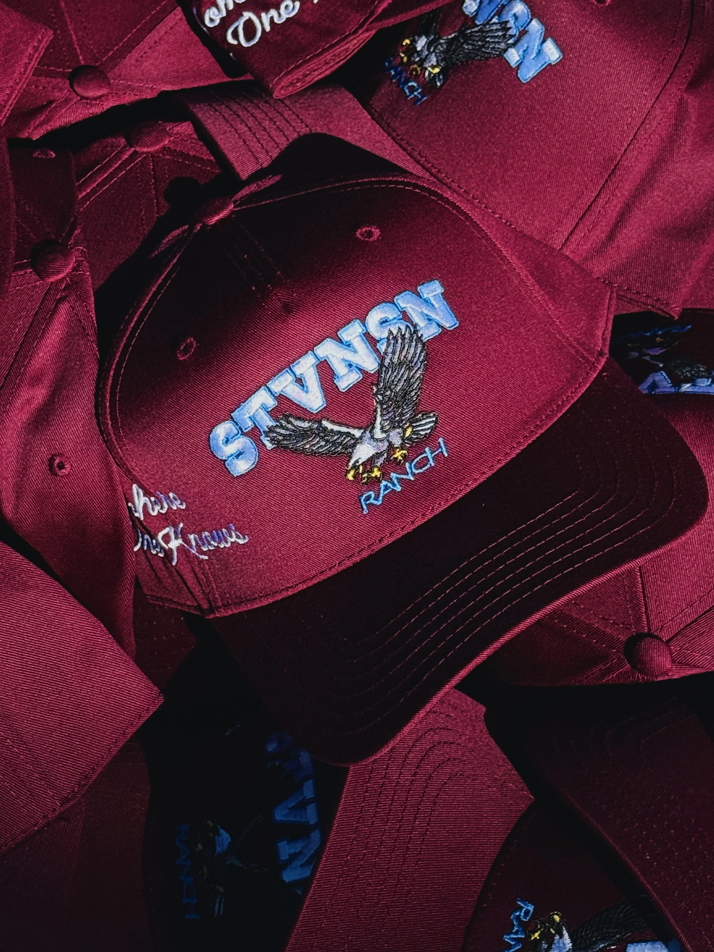 Red baseball cap featuring an embroidered eagle with outstretched wings, the word "STATES" in blue letters, and the phrase "God Bless America" embroidered in white cursive. The cap is decorated with matching red stitching and buttons.