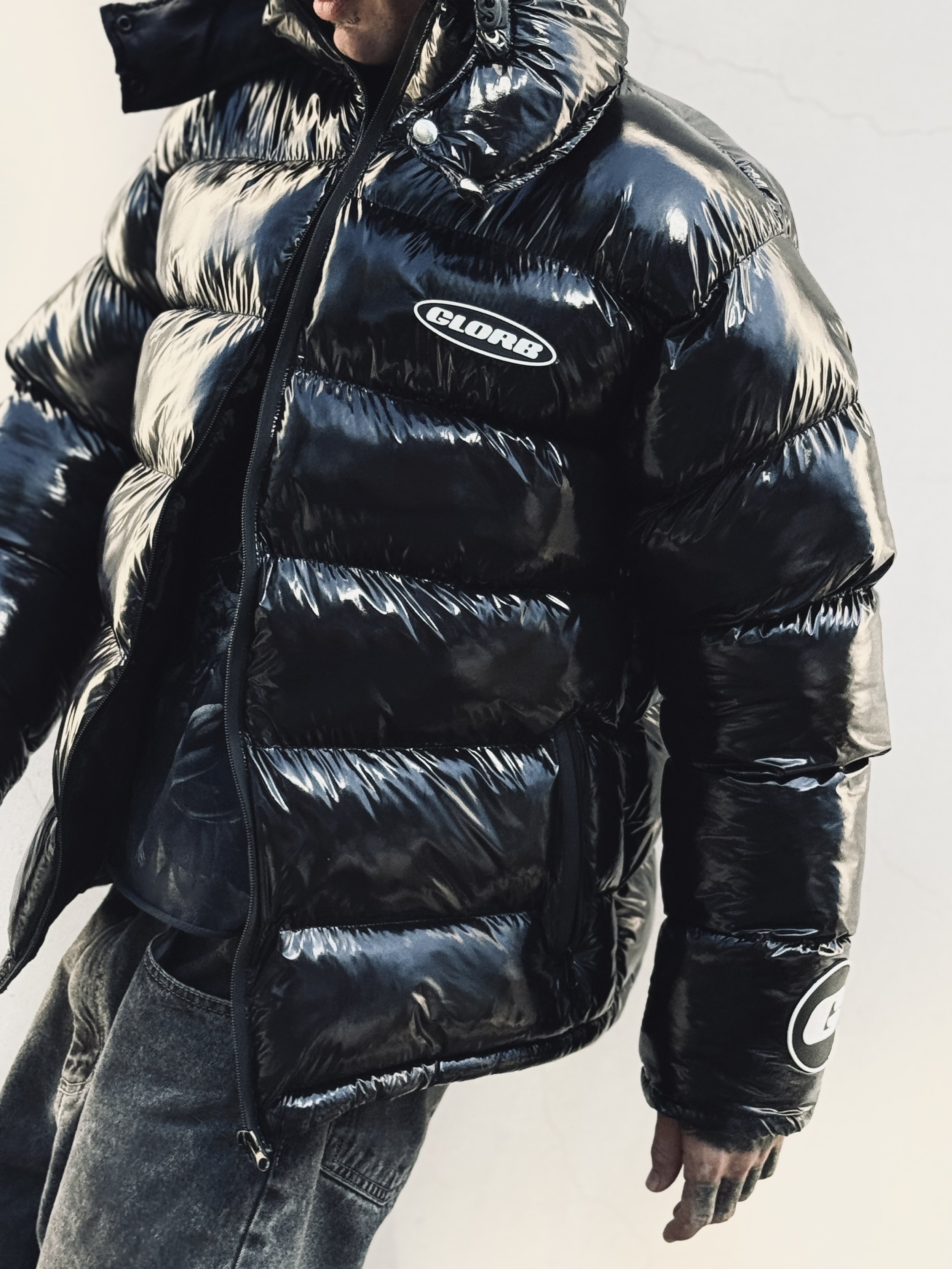 Close-up of a person wearing a shiny black puffer jacket with an embroidered patch that says 'GLOBE'. The individual has their hand relaxed by their side and is wearing gray jeans and a black Nike athletic sleeve.