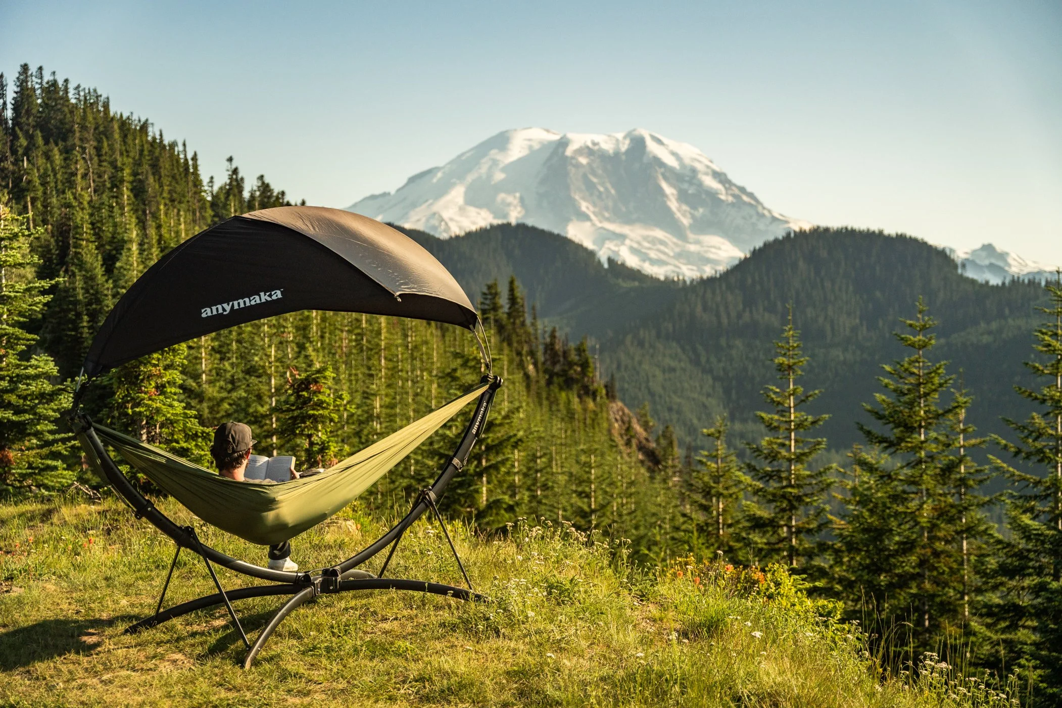 Person relaxing in a hammock beneath a large umbrella, surrounded by green trees with snow-capped mountains in the background.