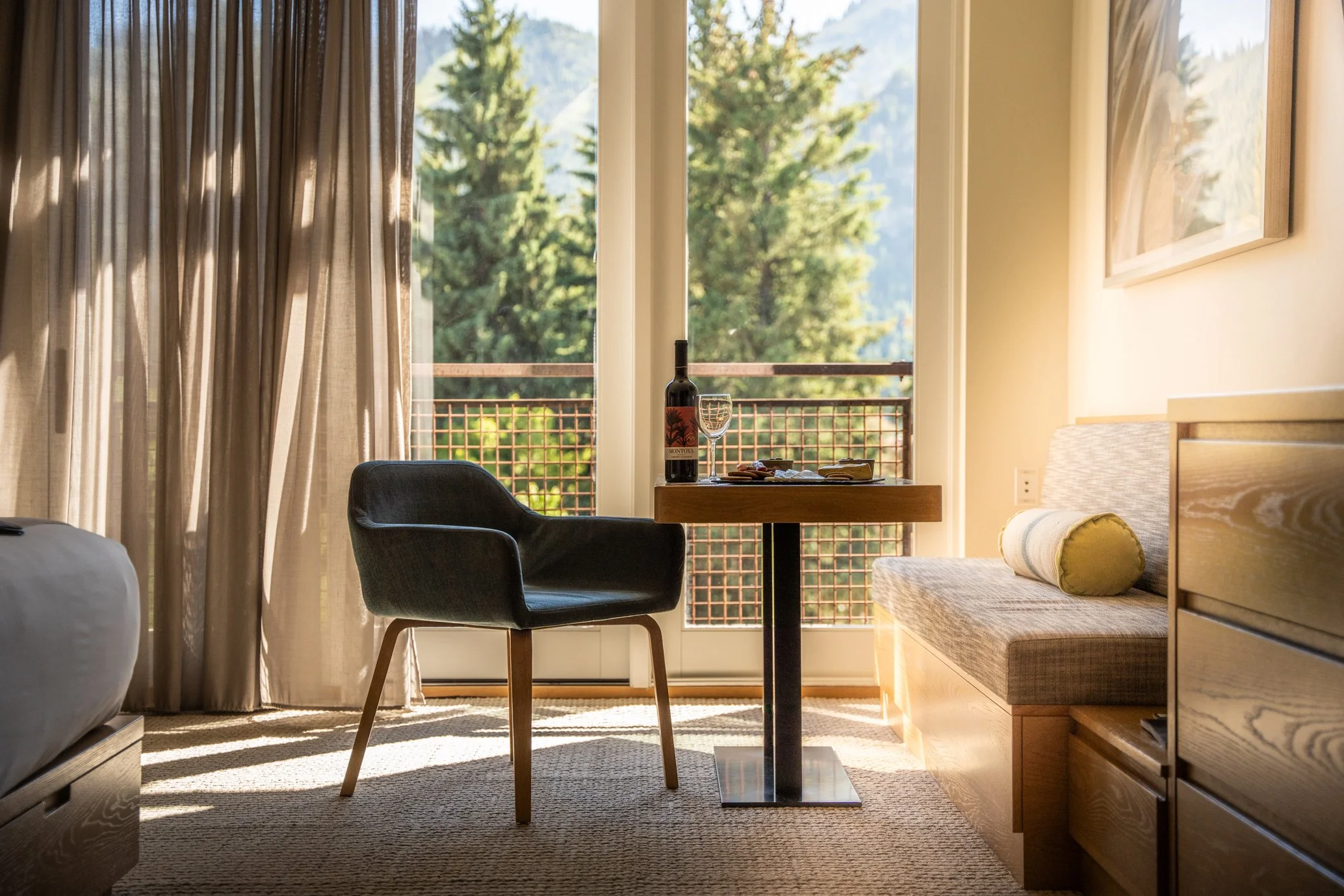 Hotel room with a window view, a black armchair, a small table with a wine bottle and glass, a nearby bench with a pillow, beige curtains, and a wooden dresser.