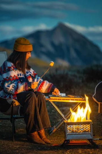 Woman sitting outdoors at night near a campfire, roasting marshmallows, with mountains in the background.