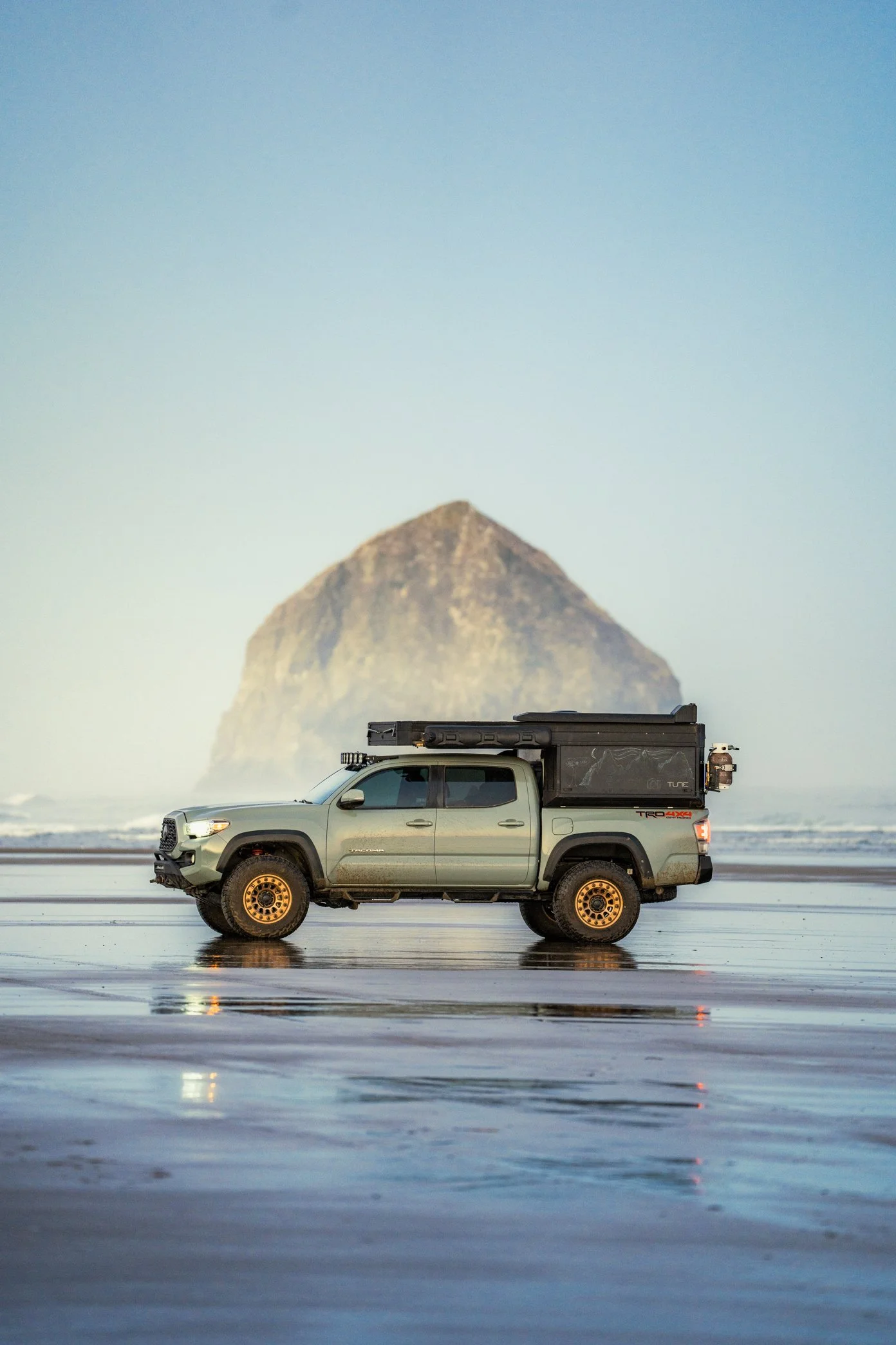 A parked pickup truck on a beach with a large rock formation in the background and a partly cloudy sky.