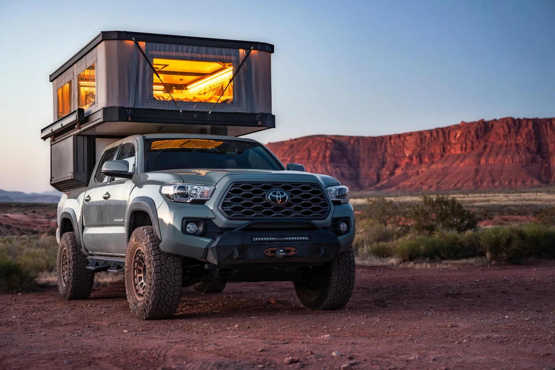 A silver Toyota Tacoma truck with off-road tires and a mounted camper in a desert landscape with red rock formations in the background at sunset.