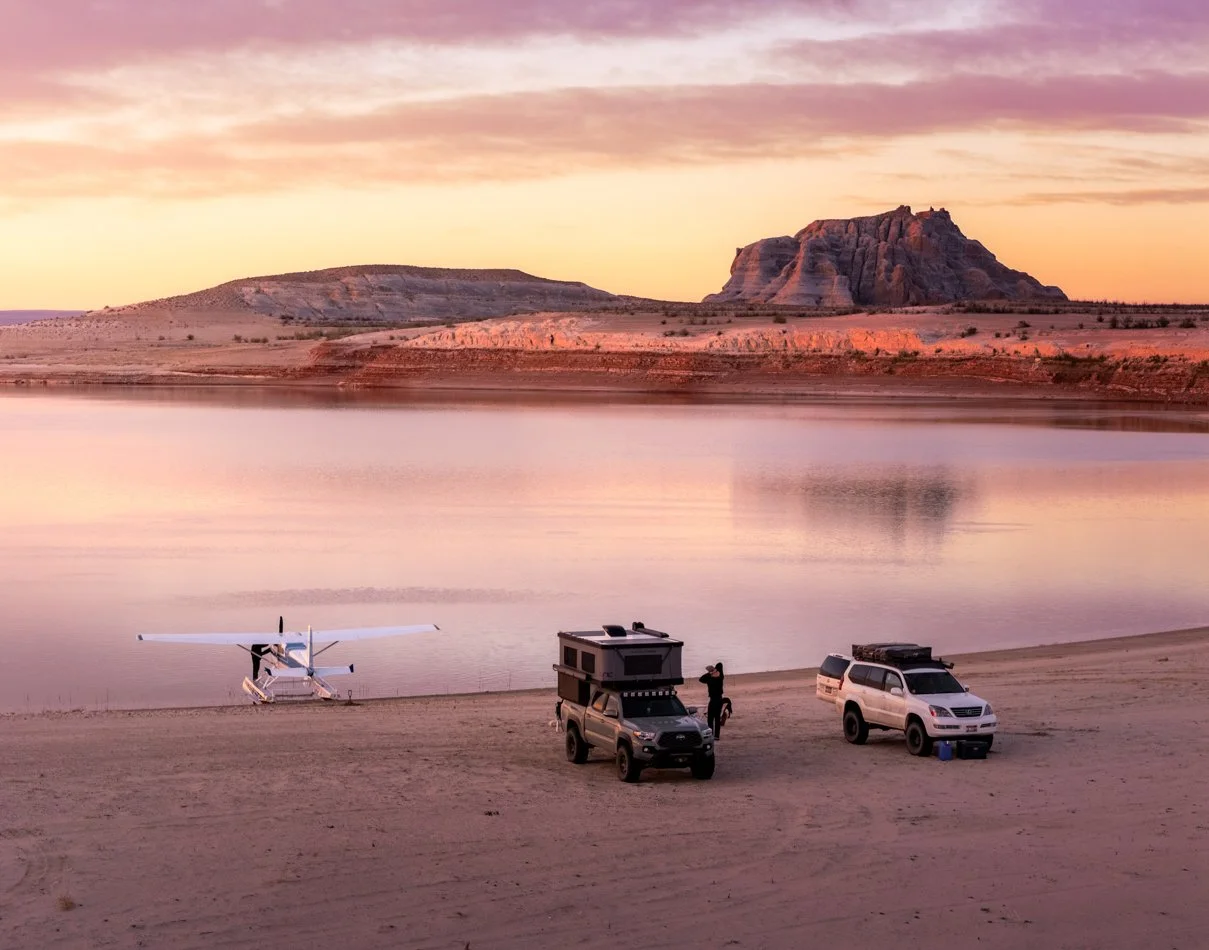 A sunset view of a river with desert mountains in the background. In the foreground, there are two off-road vehicles, one with a rooftop tent, and a person standing beside one of the vehicles. A small seaplane is parked on the sandy shore near the wa