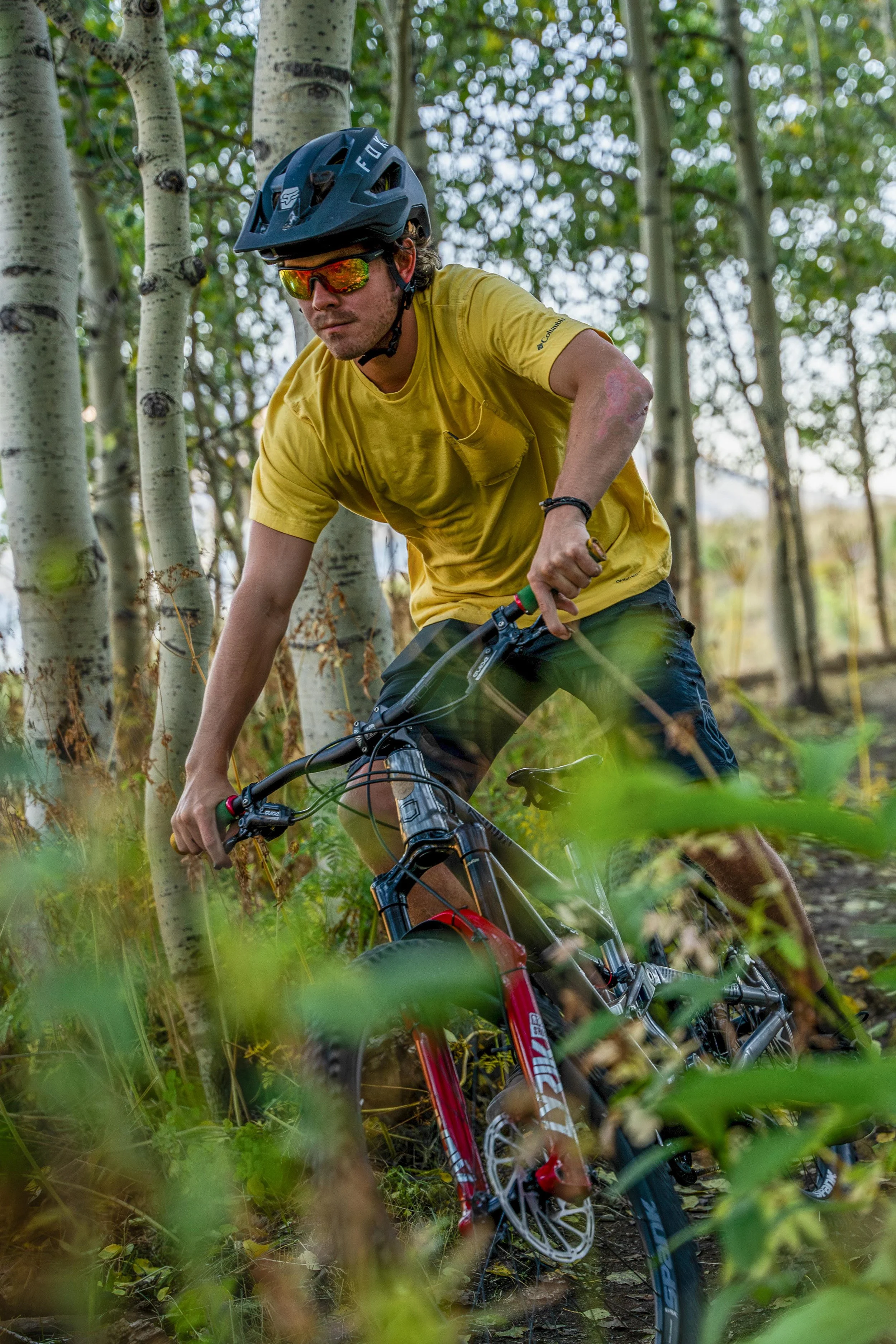 A man mountain biking through a forested trail, wearing a yellow t-shirt, helmet, and sunglasses.
