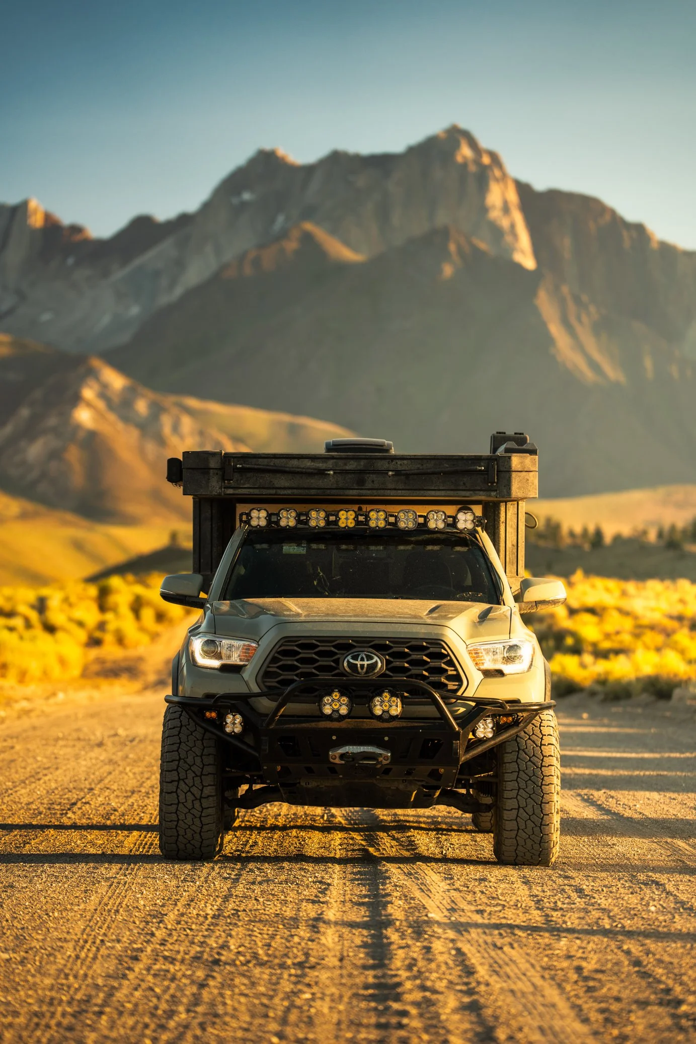 Front of a Toyota pickup truck equipped for off-road with large tires, bumper, and multiple lights, parked on a dirt road with mountainous landscape in the background during sunset.