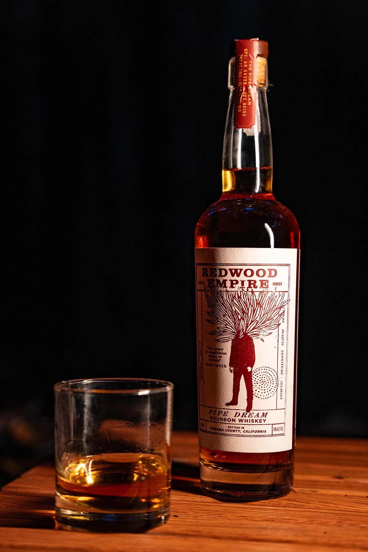 A bottle of Redwood Empire Pipe Dream Bourbon Whiskey and a glass of whiskey on a wooden surface with a dark background.