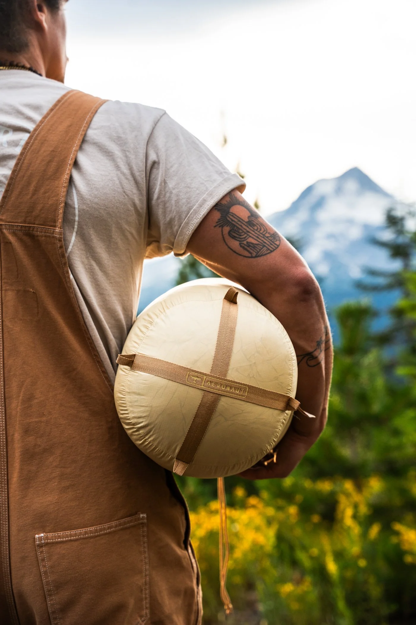 Close-up of a person wearing a beige t-shirt and brown vest, holding a yellow round tent or sleeping bag with an adjustable strap, against a backdrop of mountains and greenery.