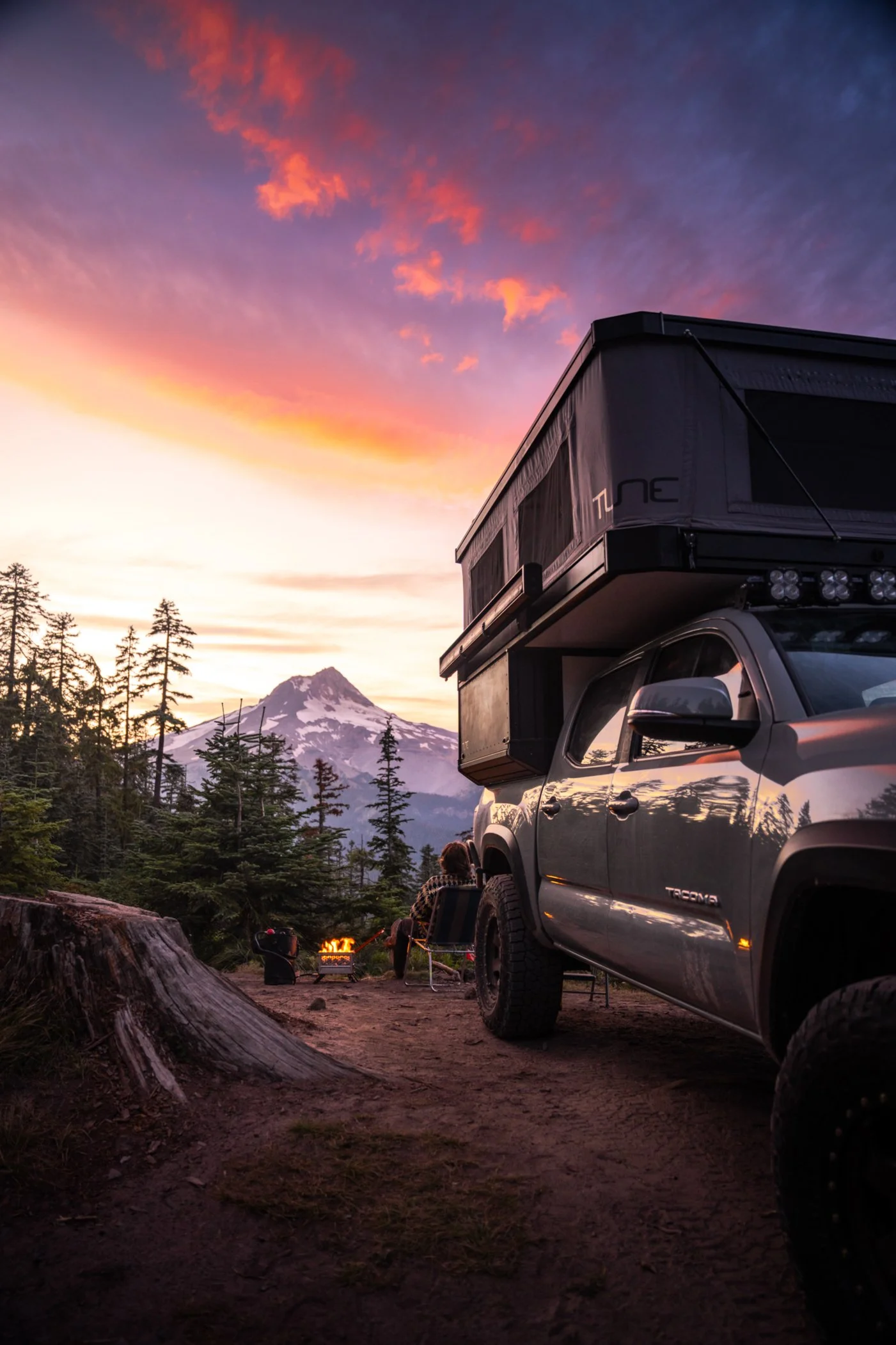 A camper truck parked near a campfire in a forested area with Mount Hood and a colorful sunset in the background.