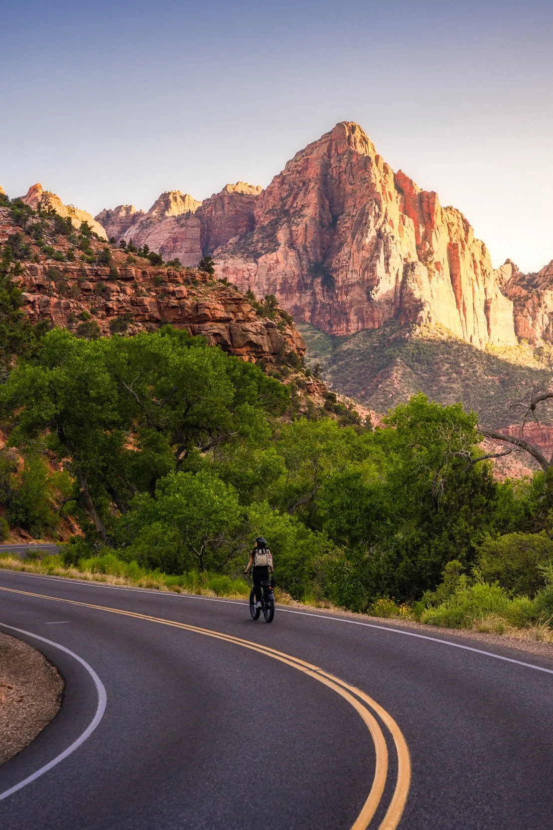 A person riding a bicycle on a winding road in a mountainous area with lush green trees and large red rock formations in the background, under a clear sky.