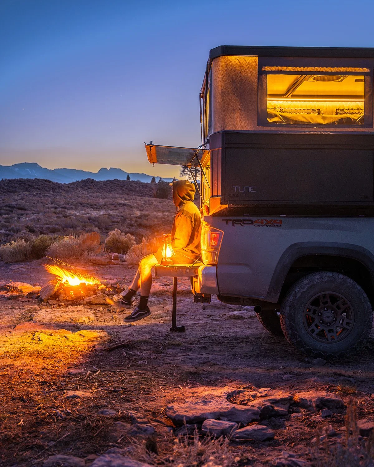 Person in yellow hoodie sitting on the tailgate of a camper truck next to a campfire at sunset in a desert landscape.