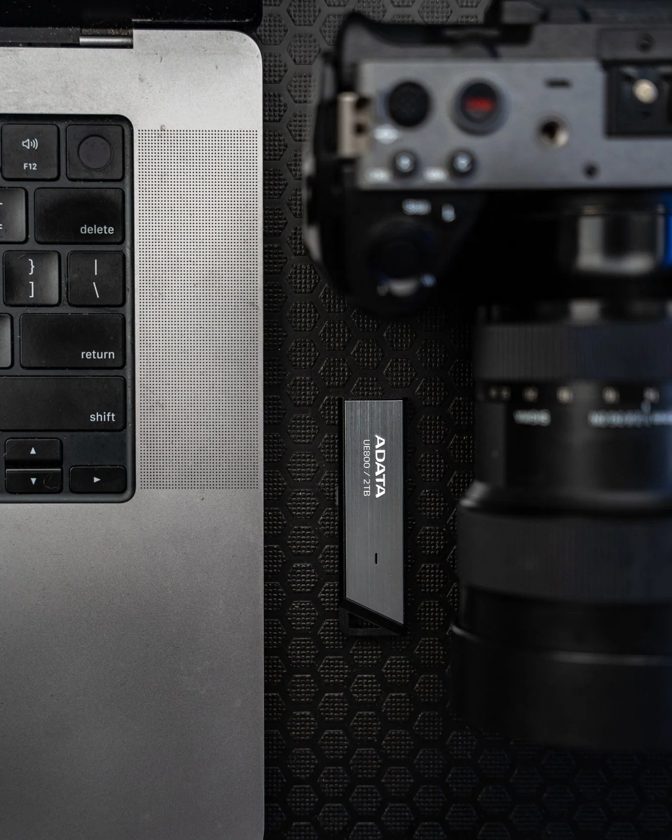 Top view of a laptop keyboard, a black camera, and an ADATA external solid-state drive on a black honeycomb textured surface.