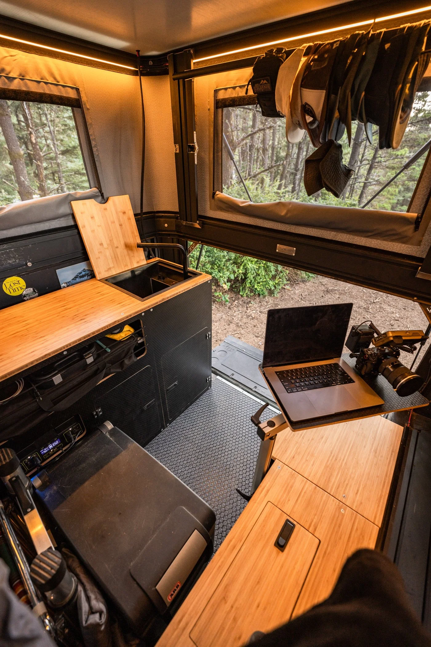 Interior of a compact camper van or truck converted into a living space with wooden countertops, a small sink, a laptop, a camera, and hanging hats and gloves, with a forest view outside the windows.