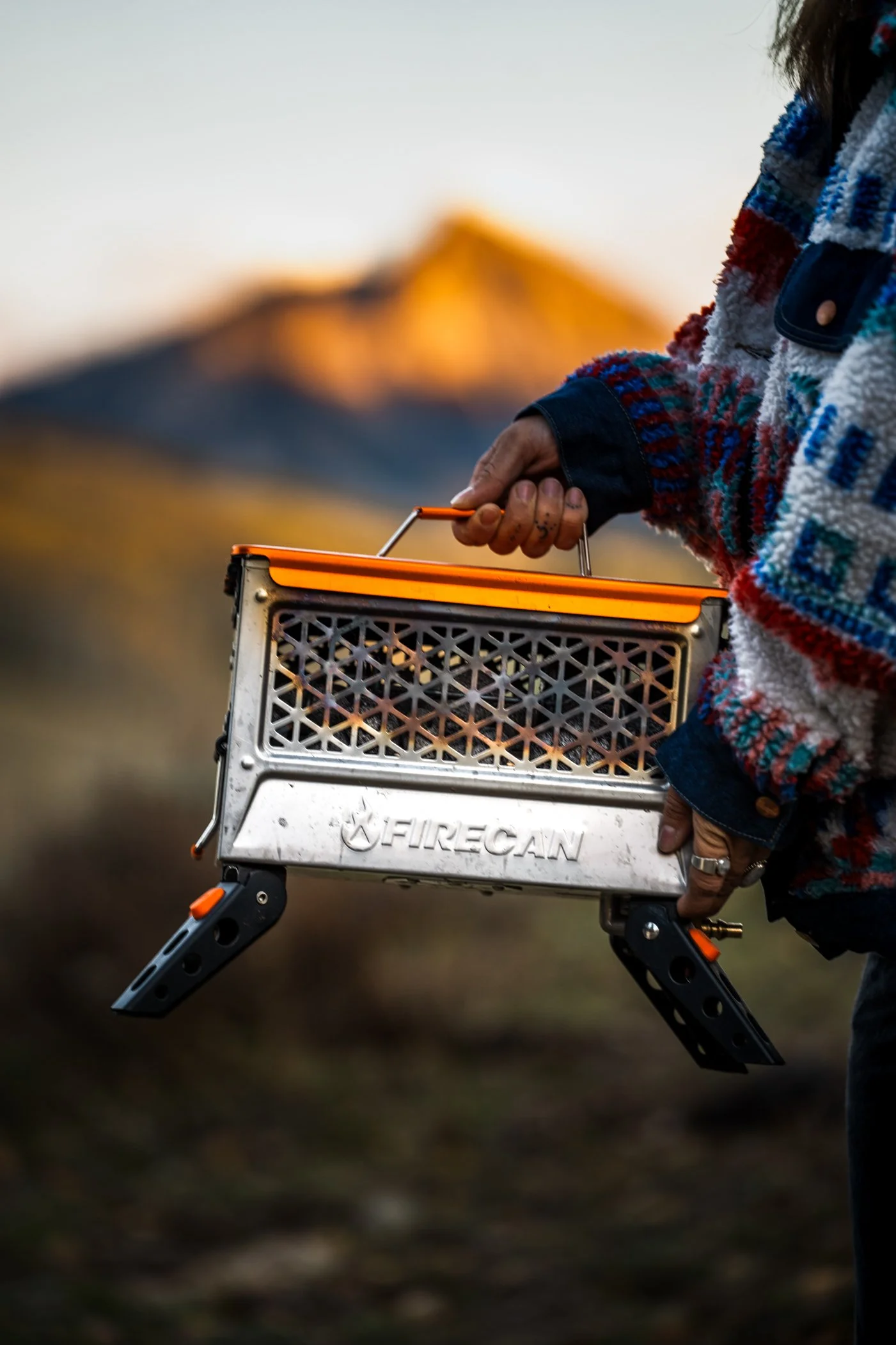 Person holding a portable camping stove outdoors with mountains in the background during sunset.