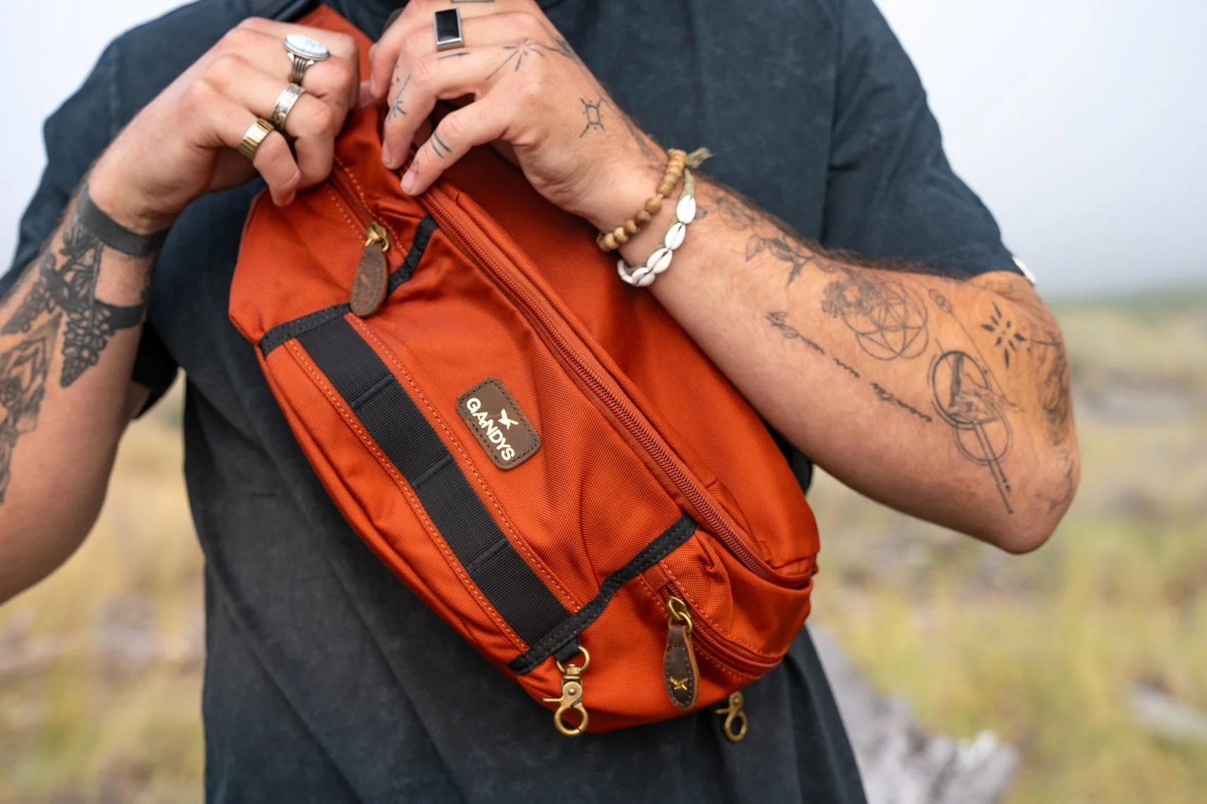 Person adjusting an orange GANDYS brand sling bag outdoors, wearing multiple rings and bracelets, with tattoos on their arm.
