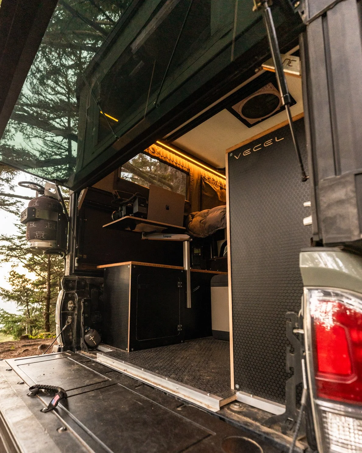 Rear view of a camper van with a view into the interior, showing a small desk with a laptop, storage cabinets, a bed with bedding, and interior lighting, parked in a wooded area.