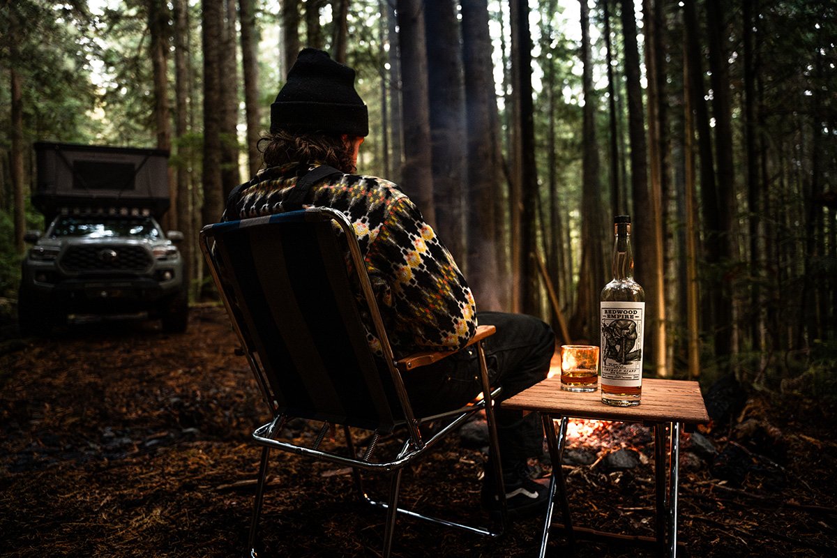 A person sitting in a camping chair in a forest, with a bottle of alcohol and a glass on a small table in front of them, and a vehicle in the background.