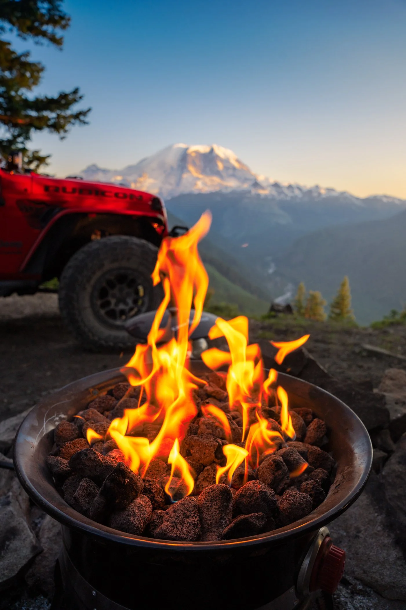 A campfire with flames burning over lava rocks in the foreground, with a red off-road vehicle and a mountain in the background, possibly Mount Rainier, during sunset.