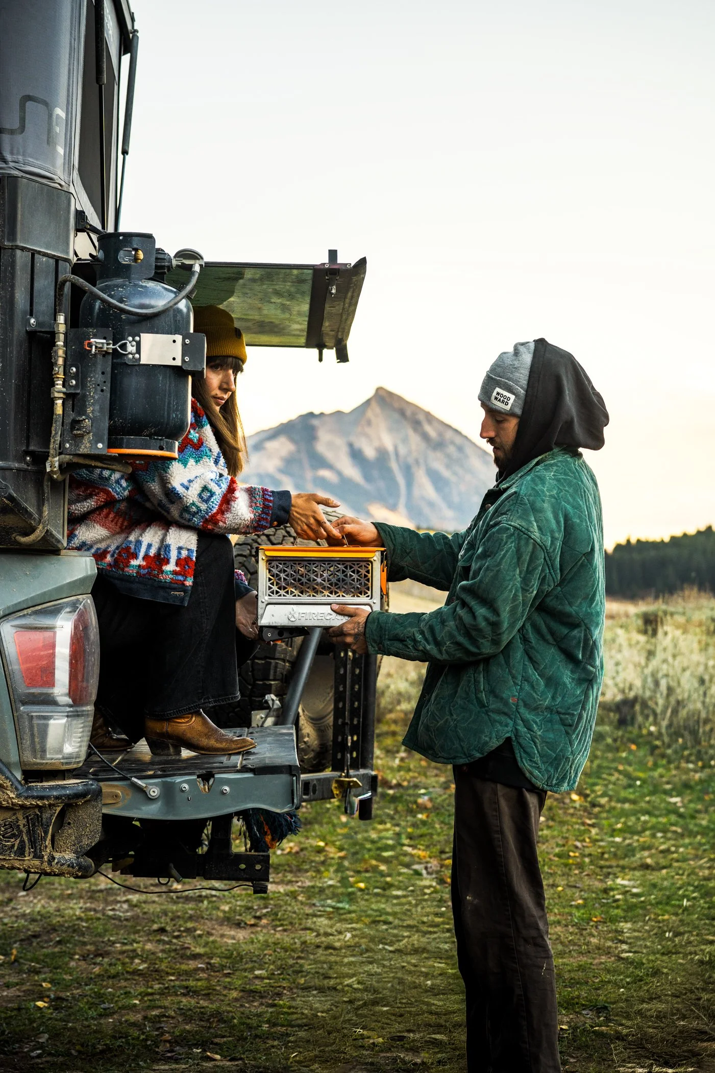 Two people exchanging gear from the back of a truck outdoors with a mountain in the background, during sunset.