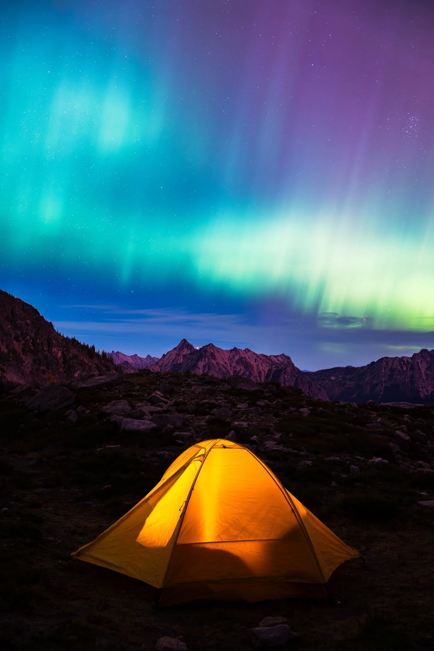 A glowing yellow tent in a mountain landscape at night with the northern lights visible in the sky.
