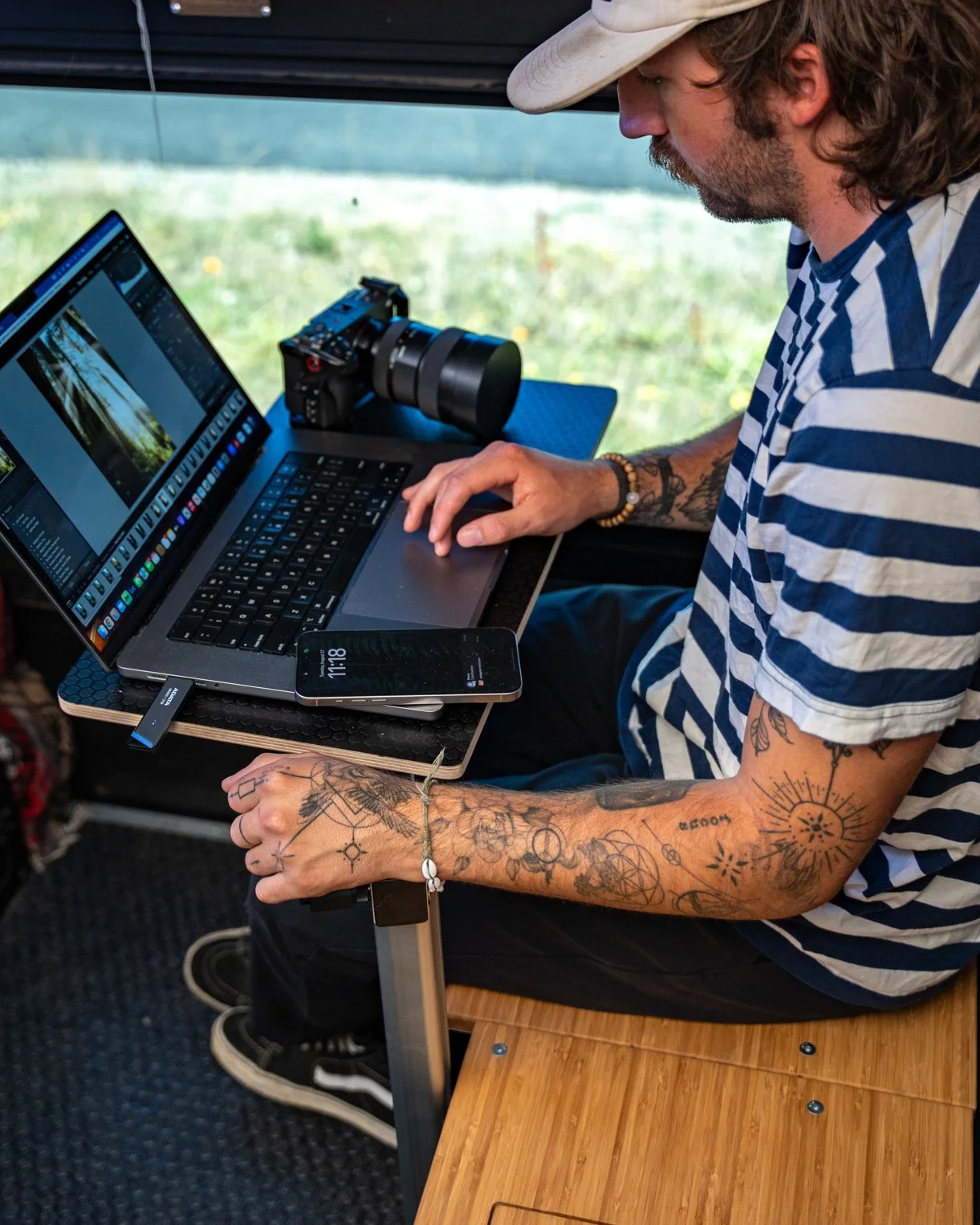 A man with tattoos on his arms, wearing a striped shirt and a hat, working on a laptop. There is a camera and a smartphone on the table, with a green outdoor background visible.
