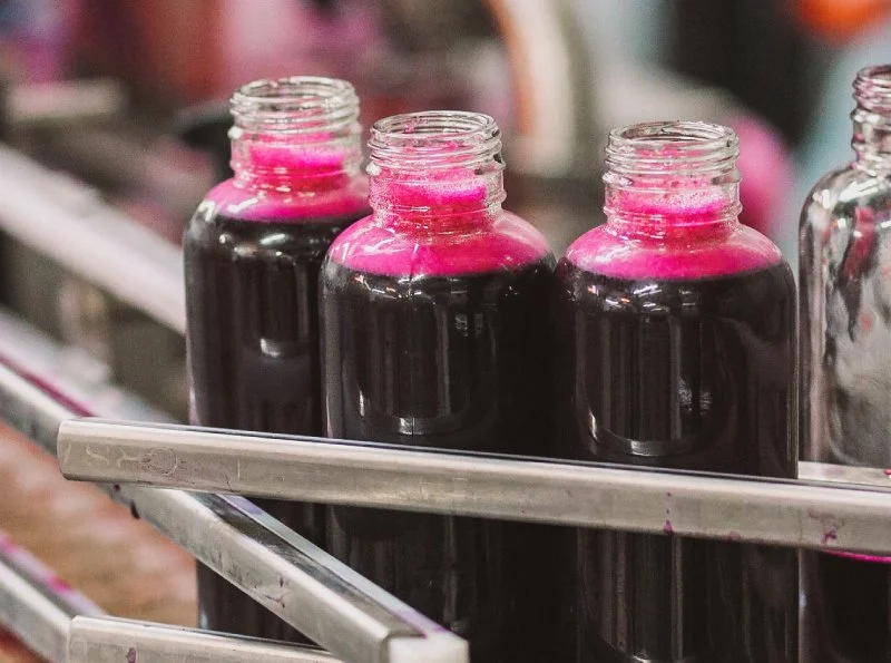 Four glass bottles filled with dark purple liquid, with pink ink or dye on the necks, on a metal shelf.