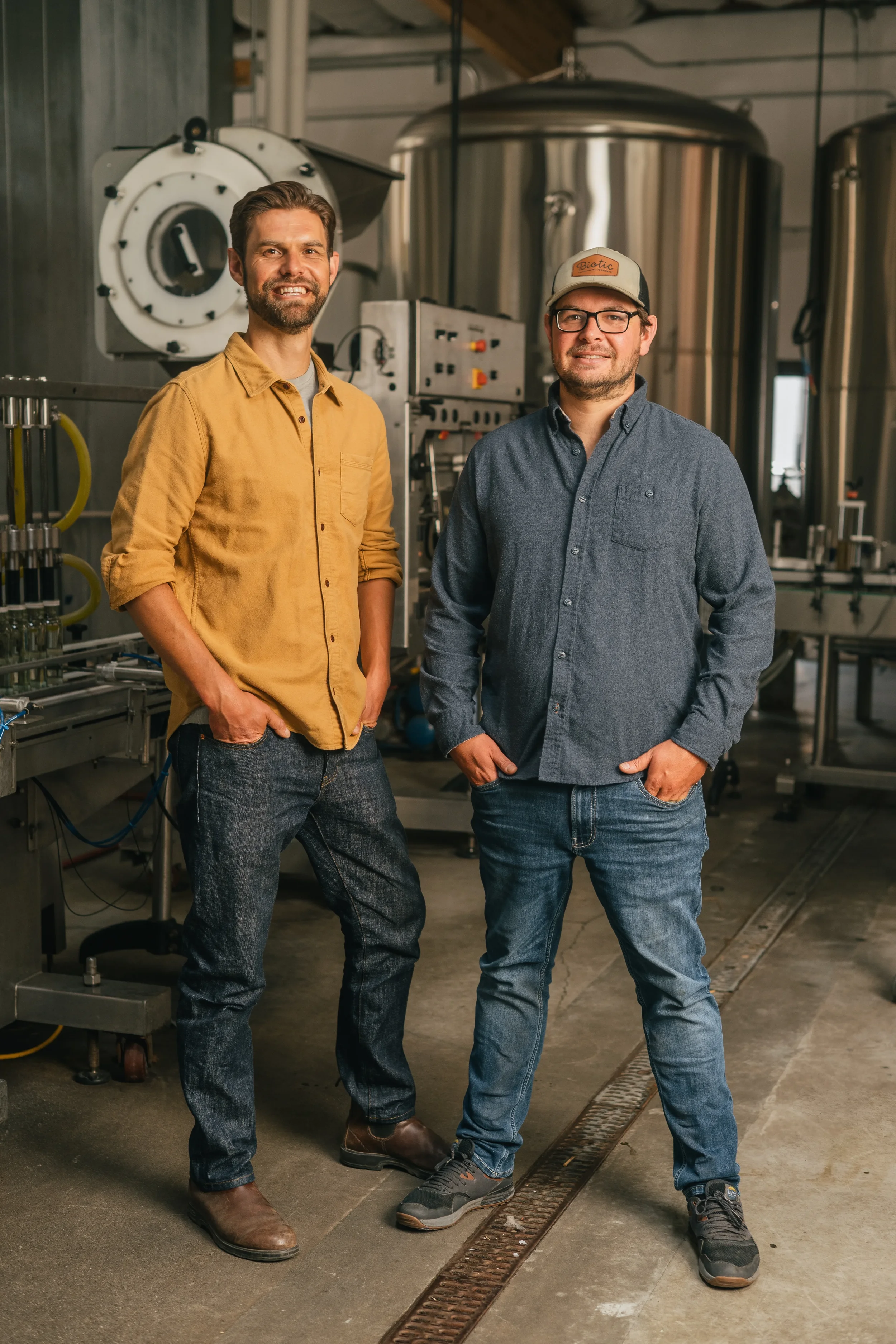 Two men standing in a brewery or industrial setting with stainless steel brewing or processing tanks in the background.