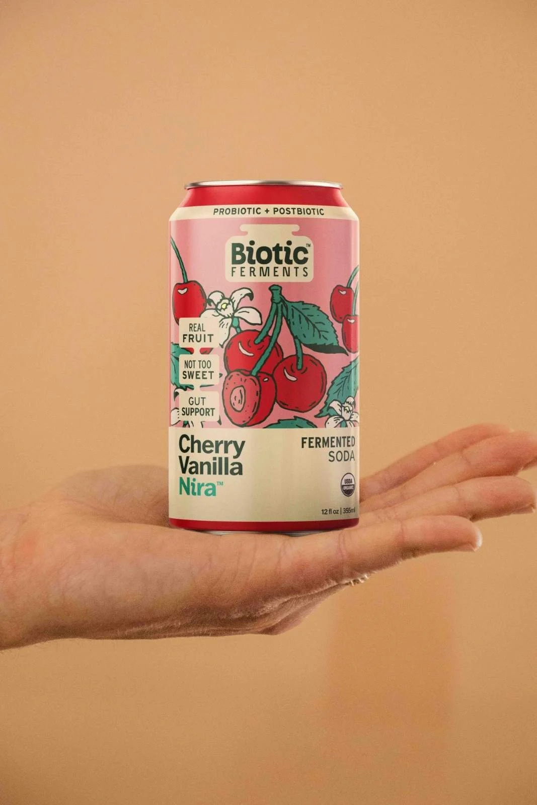 A hand holding a can of Biotic Ferments Cherry Vanilla Nira probiotic soda against a beige background.