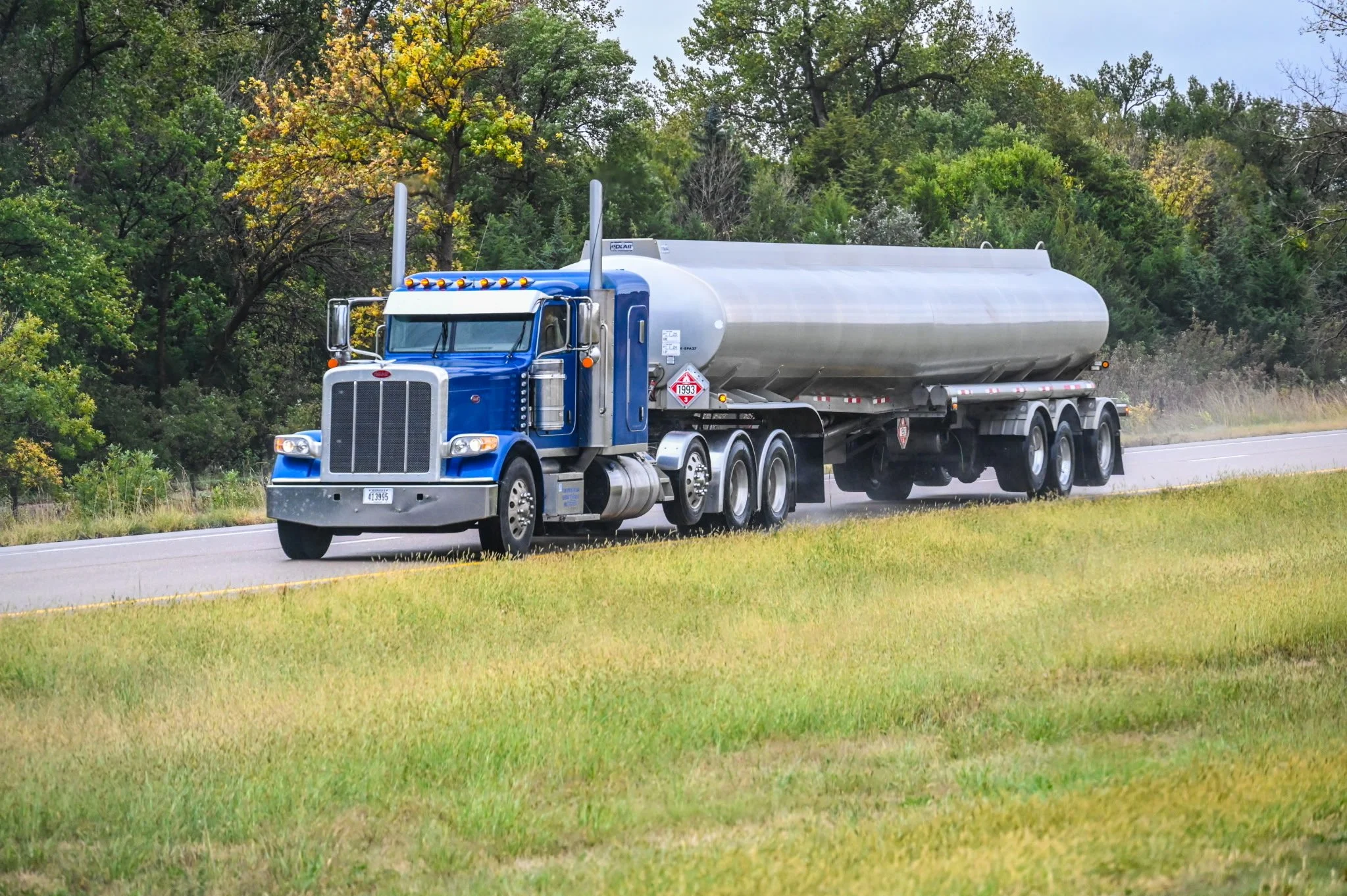 A large blue tanker semi-truck driving on a rural highway with trees and grass on the roadside.