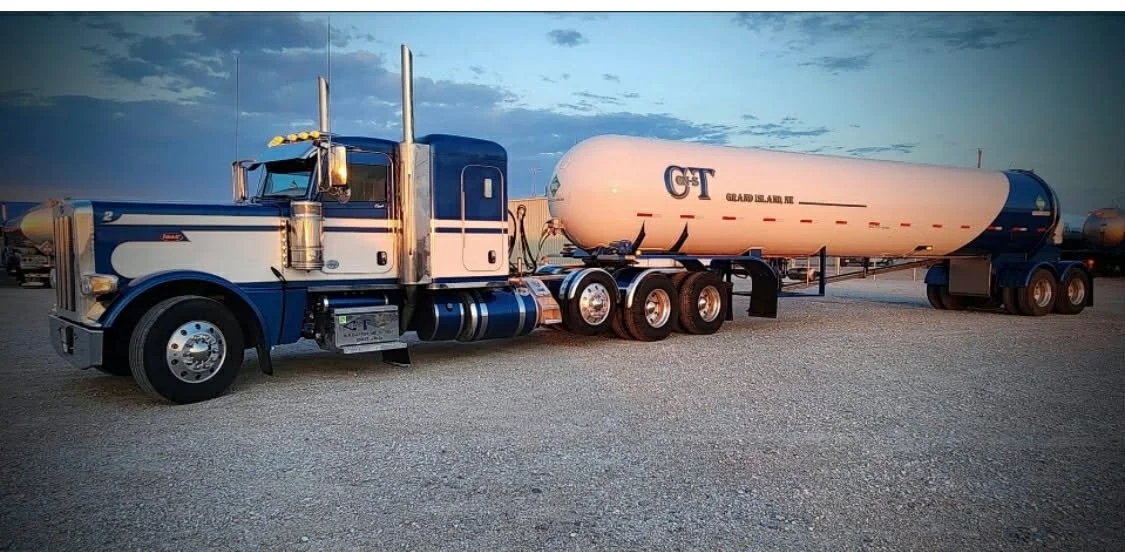 A semi-truck with a blue and white cab and a white tank trailer on a gravel lot during sunset.