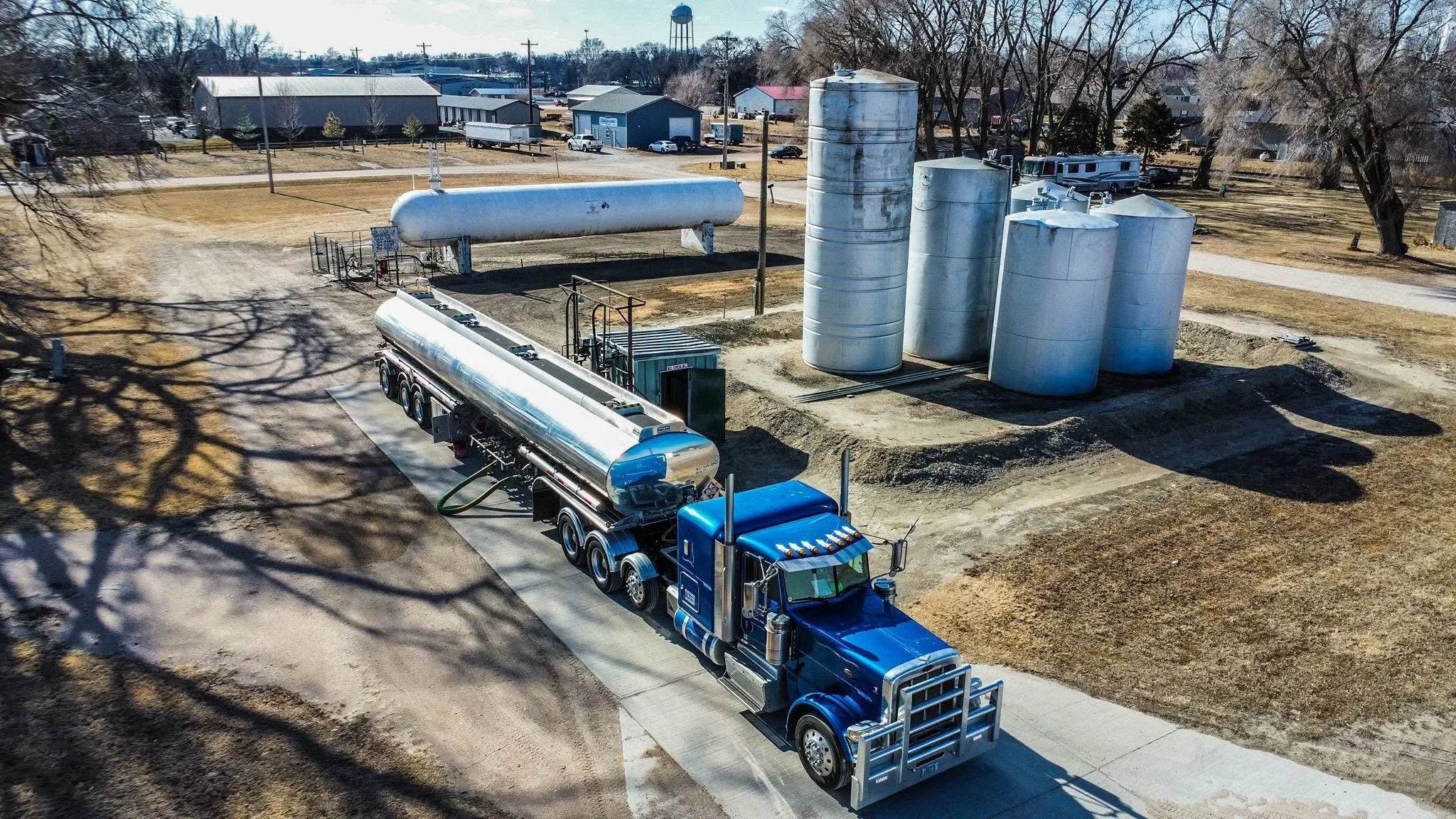 A large blue semi-truck with a shiny metal tank trailer driving on a rural road near industrial storage tanks and a water tower, with leafless trees and farm buildings in the background.