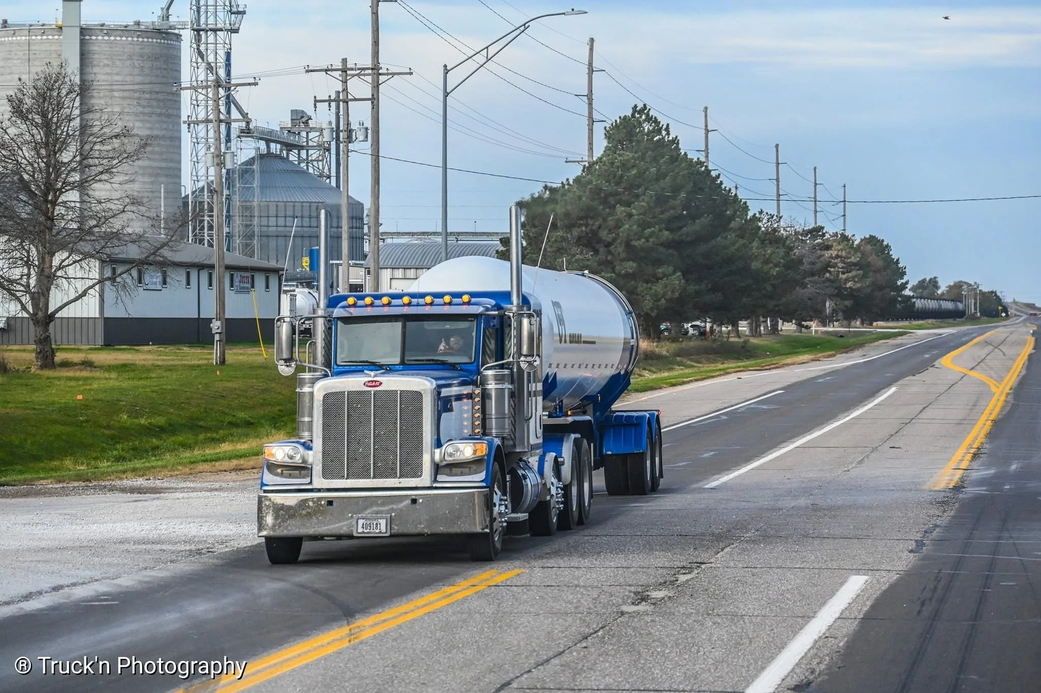 A blue semi-truck with a silver tank trailer driving on a two-lane road with yellow dividing lines, overcast sky, power lines, trees, and industrial buildings in the background.
