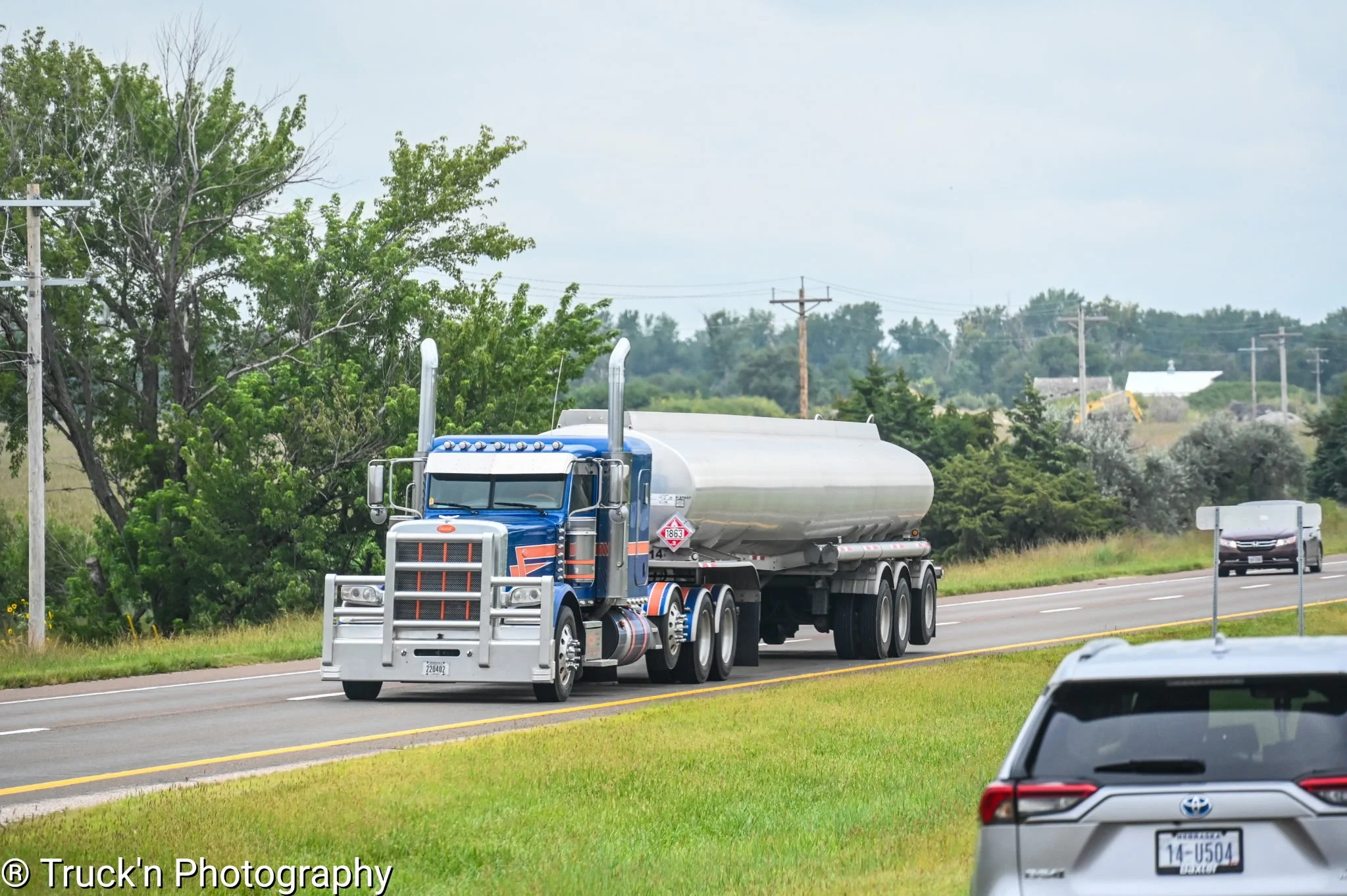 A large blue semi-truck with a silver tanker trailer driving on a highway, with greenery and power lines in the background, and another car visible in the foreground.