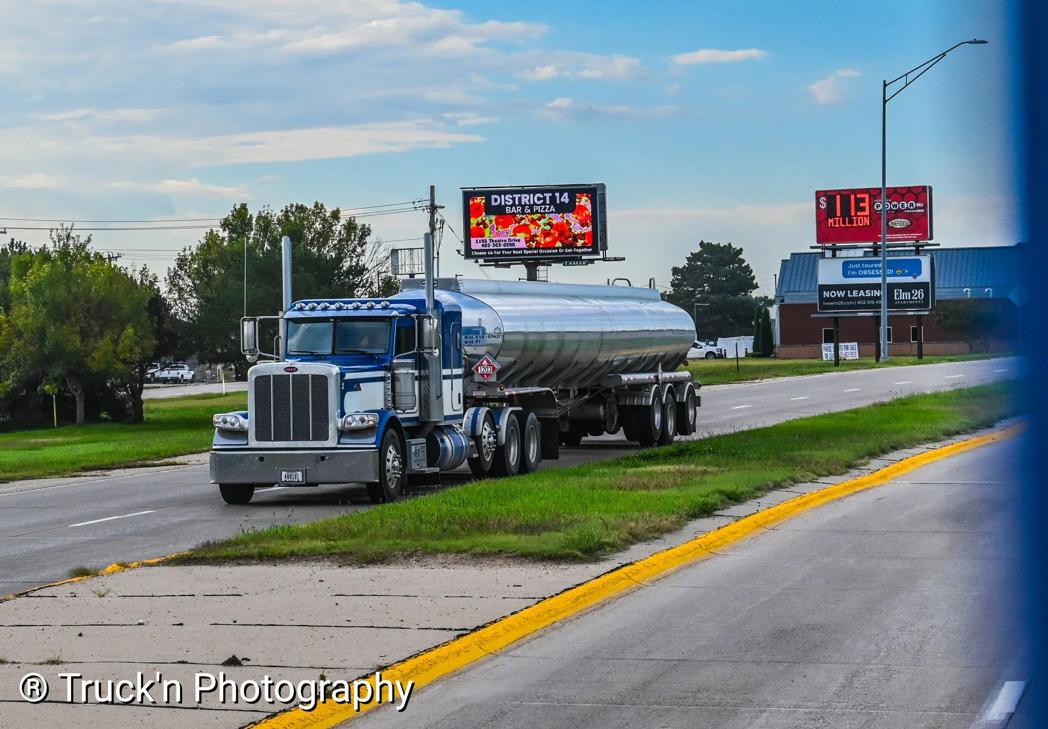 A large blue and silver tanker truck driving on a road with a grassy shoulder, under a partly cloudy sky. There are trees, billboards, and buildings in the background.