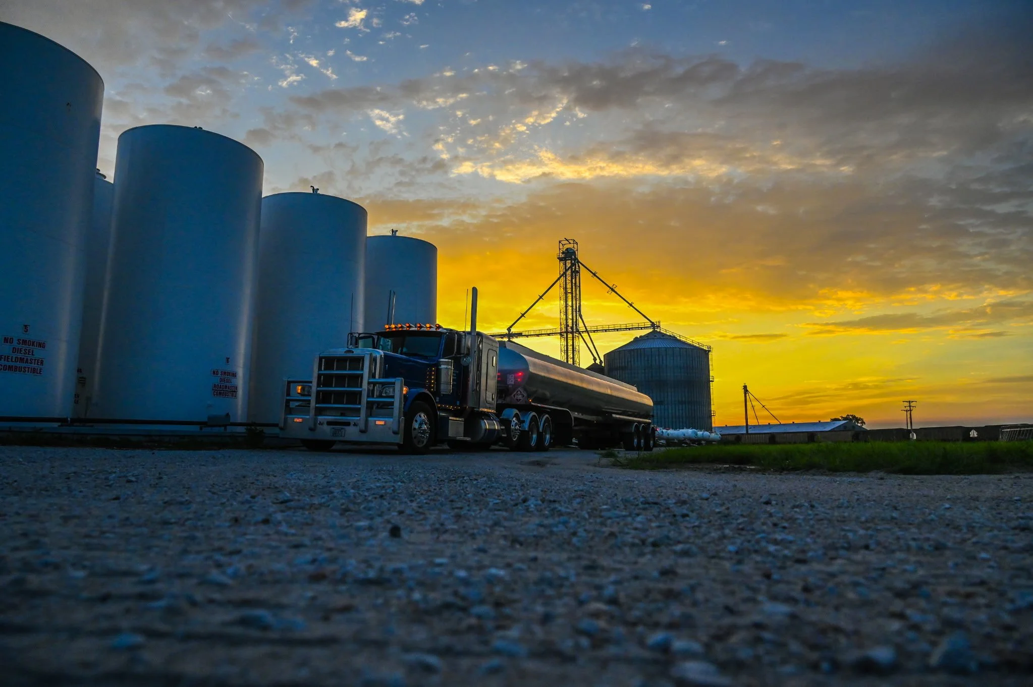 A semi-truck with a black tanker trailer parked on a gravel surface at sunset, with large white storage tanks and industrial silos in the background.