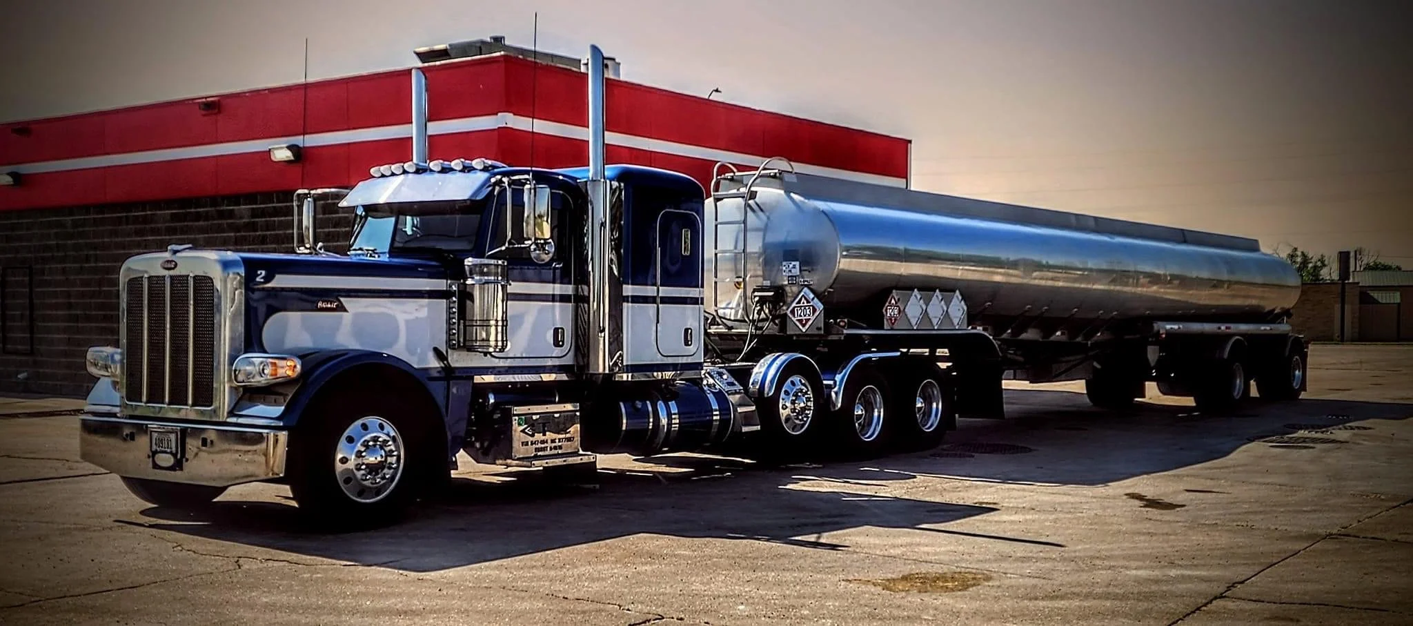 A large semi-truck with a chrome tank trailer parked on a concrete surface in front of a building with a red and black exterior.