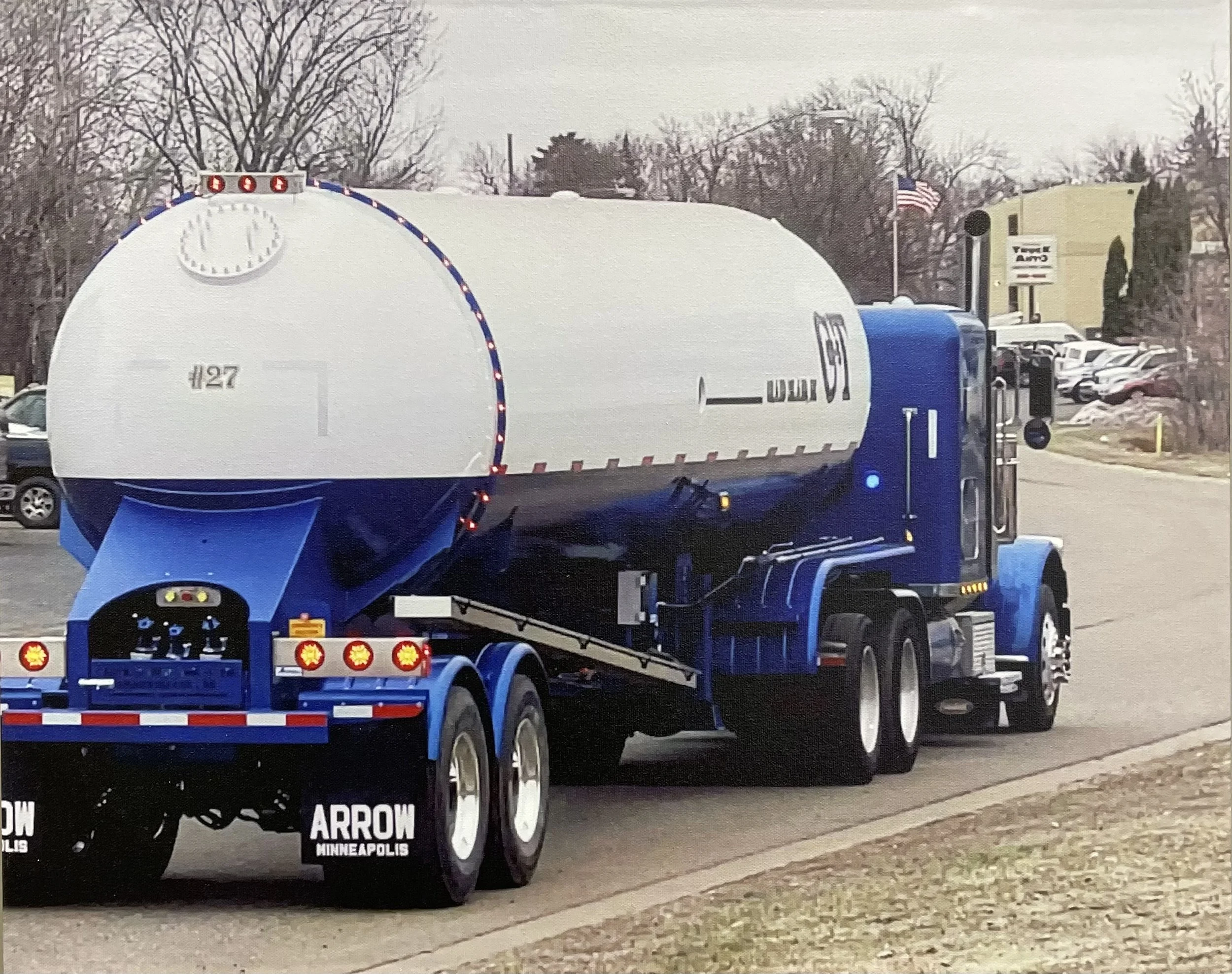 A large blue tanker truck with white tank, parked on a street, with an American flag visible in the background, and several cars parked nearby.