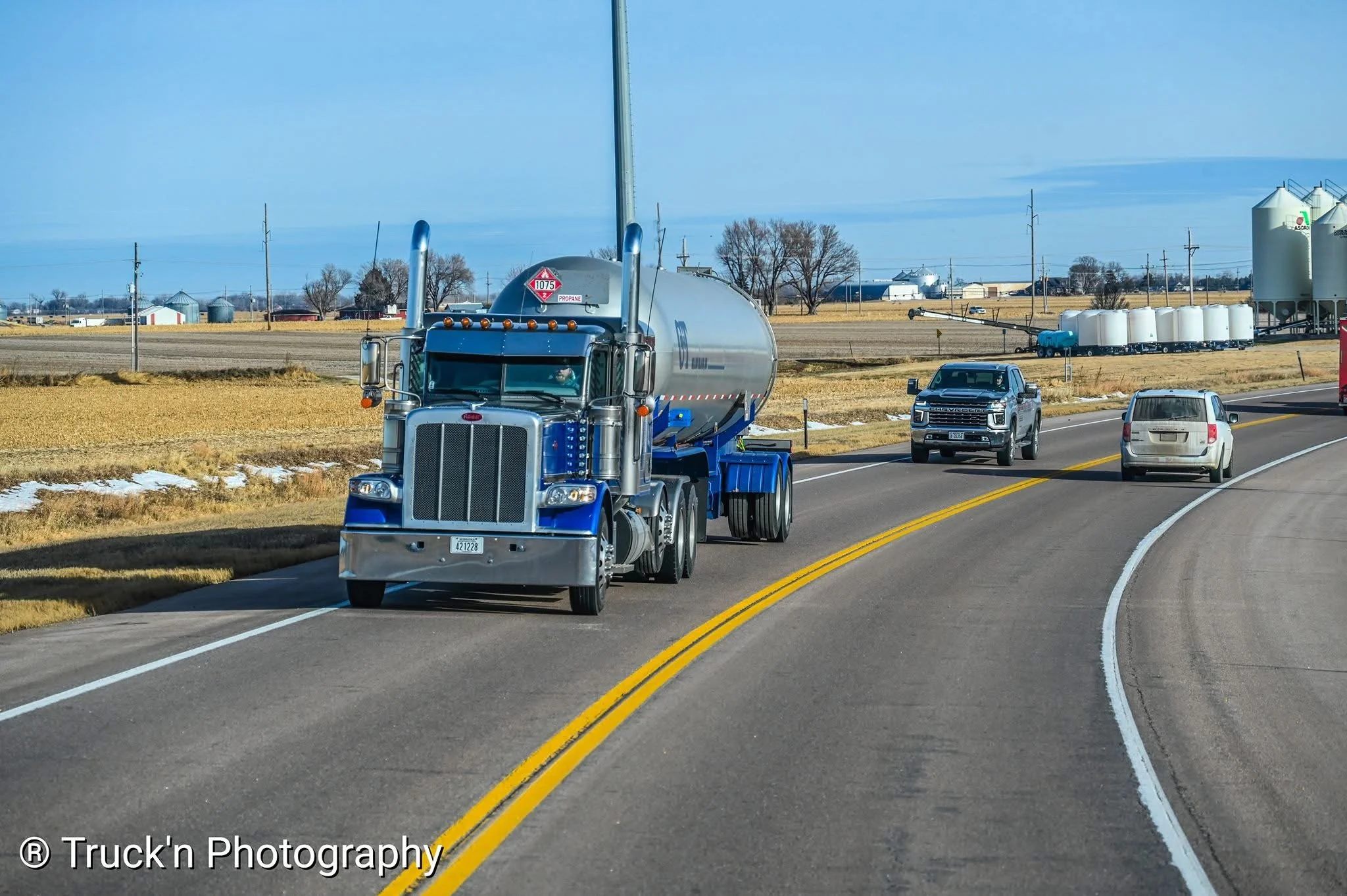 A large blue semi-truck with a chrome grille and propan logo on the tank driving on a rural road with other vehicles including a pickup truck and minivan, with fields and farm silos in the background.
