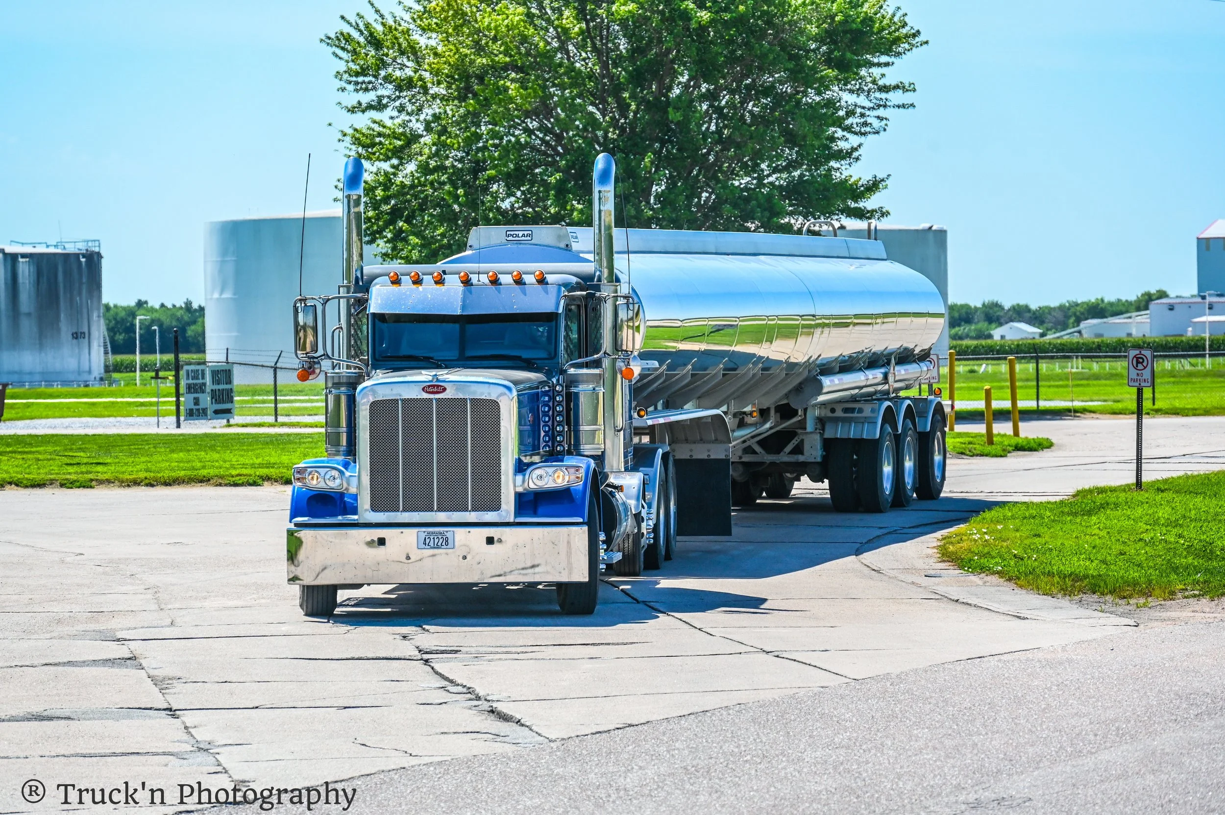 A large blue semi-truck with a shiny silver fuel tank driving on a paved road near a grassy area with industrial storage tanks and buildings in the background.