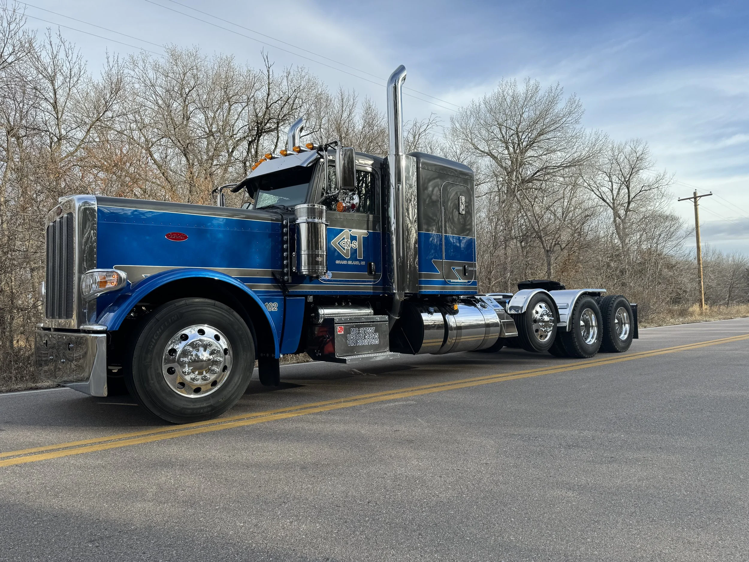 A large blue semi-truck is parked on the side of a rural road during daytime, with leafless trees and a utility pole in the background.
