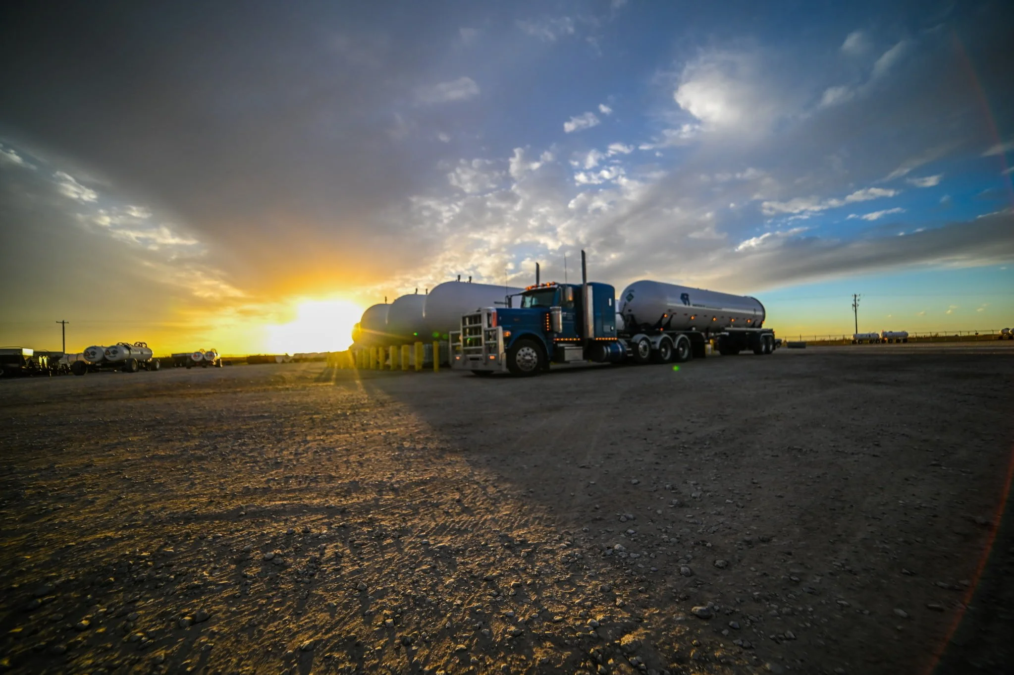 A blue semi-truck with fuel tankers along a dirt road during sunset, with a partly cloudy sky and distant vehicles.