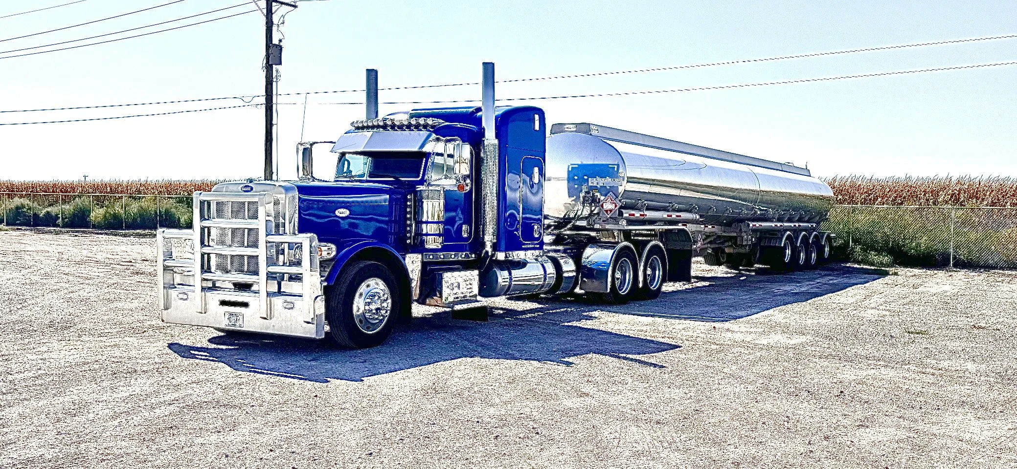A shiny blue semi-truck with chrome accents parked on a gravel lot, attached to a large cylindrical metal tanker trailer.